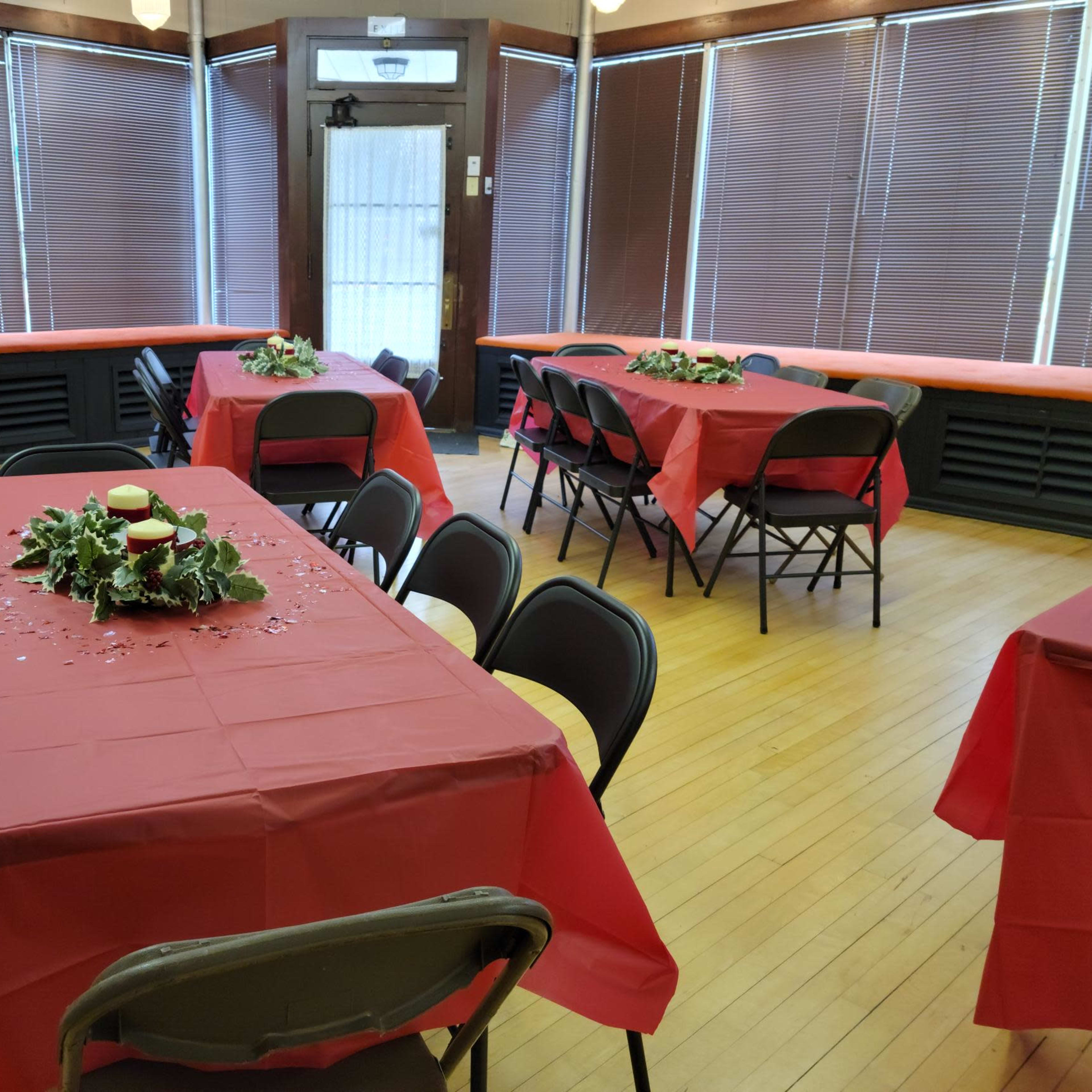 The room features several tables covered with red tablecloths, arranged with centerpieces of greenery, and is illuminated by natural light from large windows.