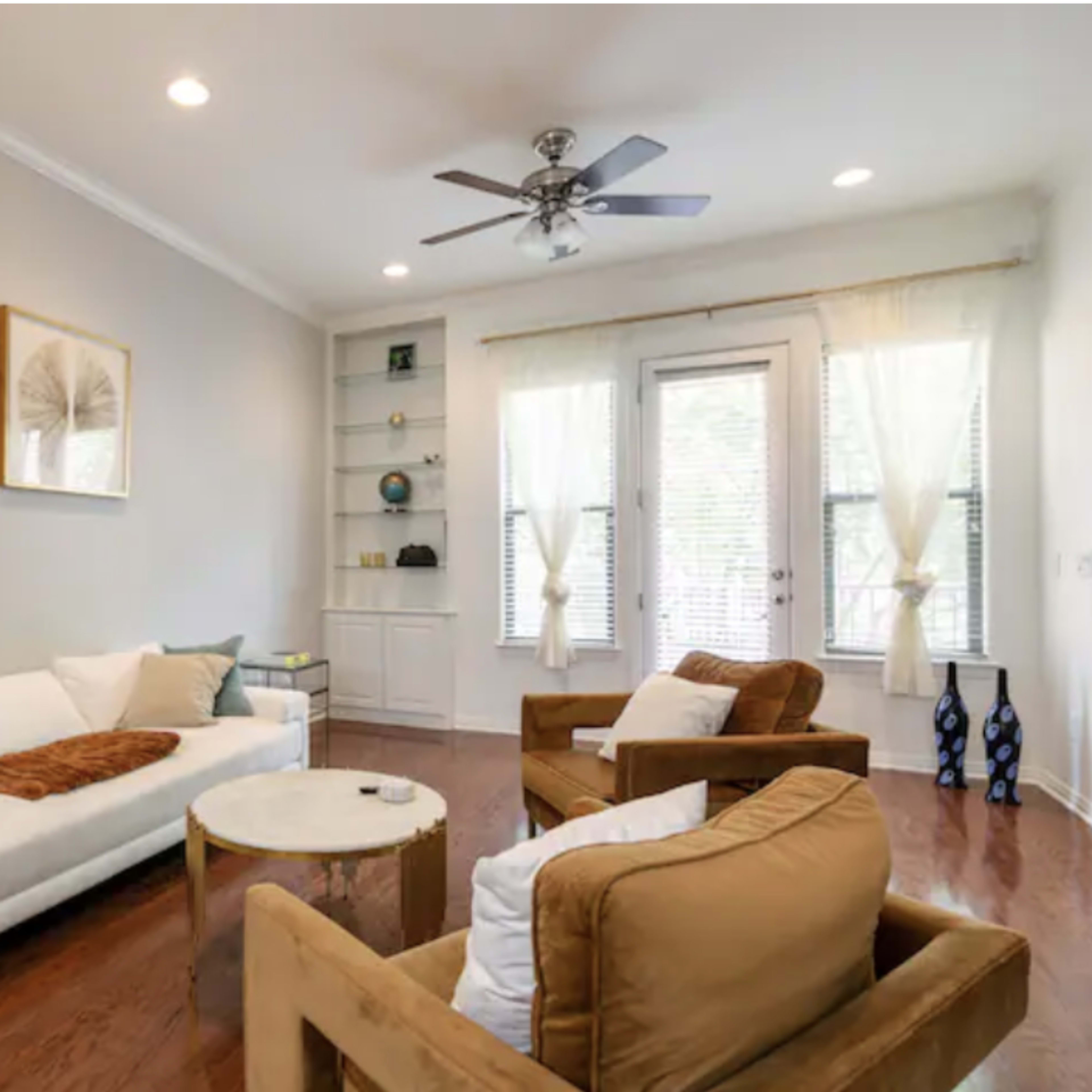 The living room features a white sofa, two brown armchairs, a round wooden coffee table, and framed art on the walls, with large windows allowing natural light.