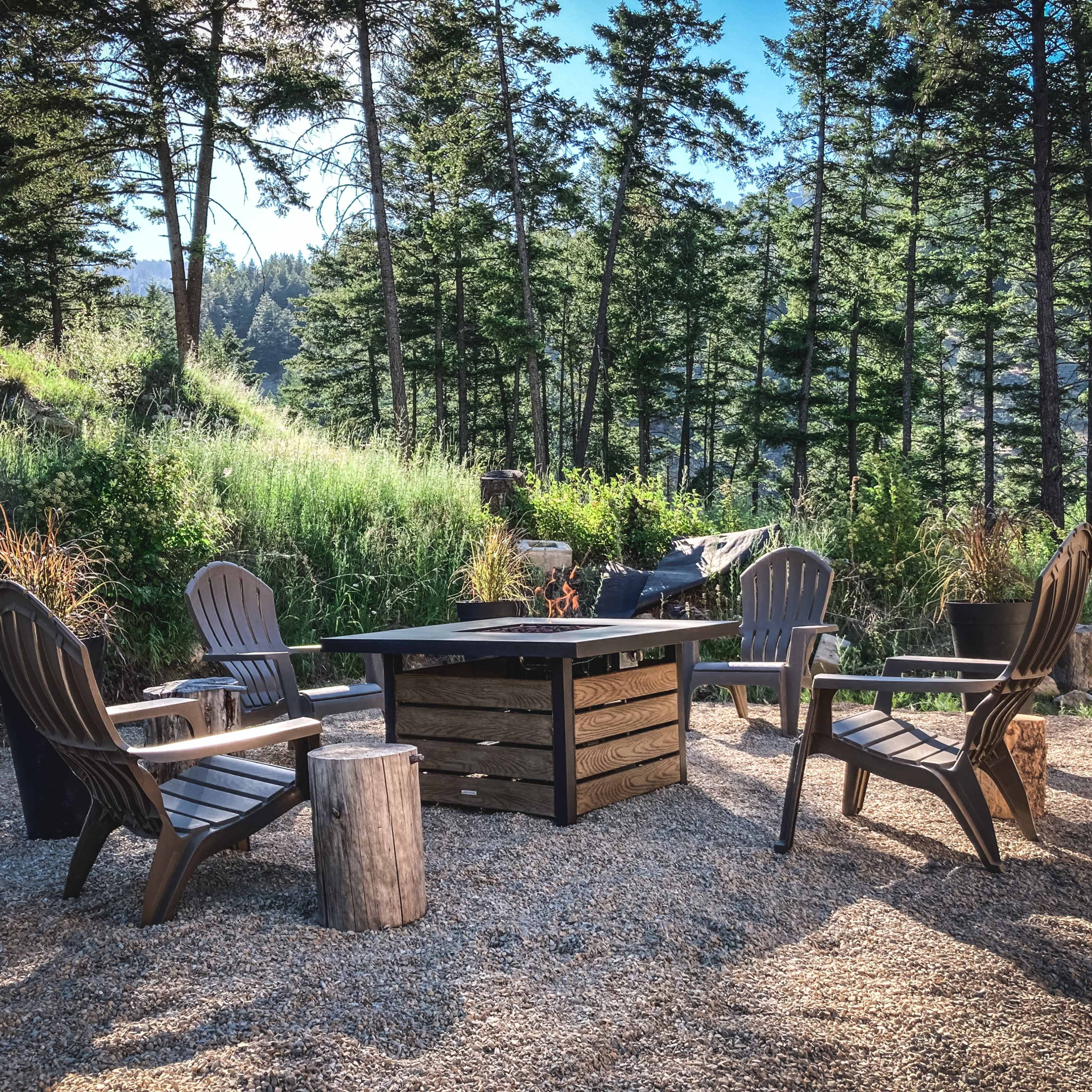 A circular seating area with four wooden chairs surrounds a rectangular fire pit, set in a gravel patch amidst a forested landscape.