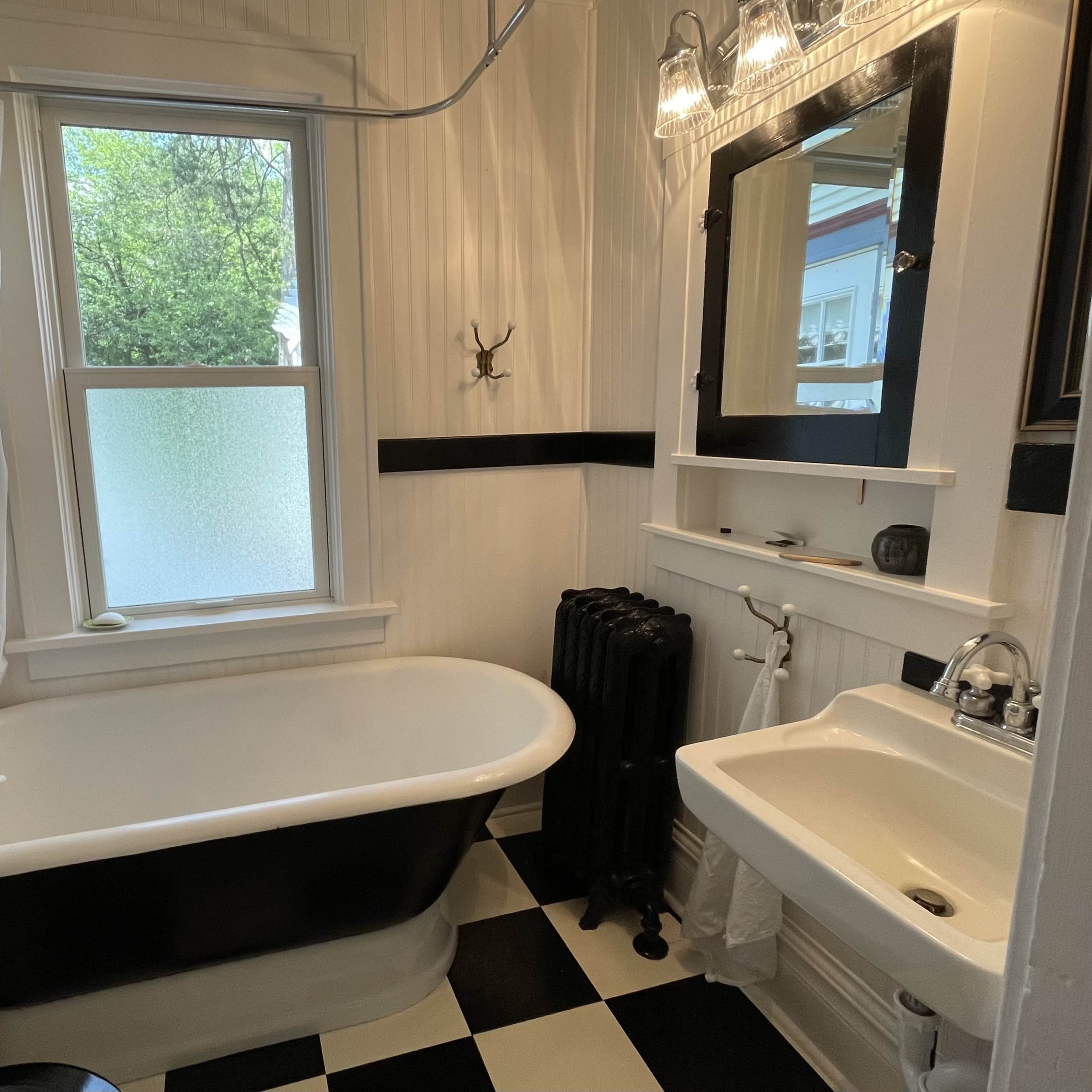The bathroom features a clawfoot bathtub, a pedestal sink, and a window with frosted glass, all set against black and white checkered flooring.