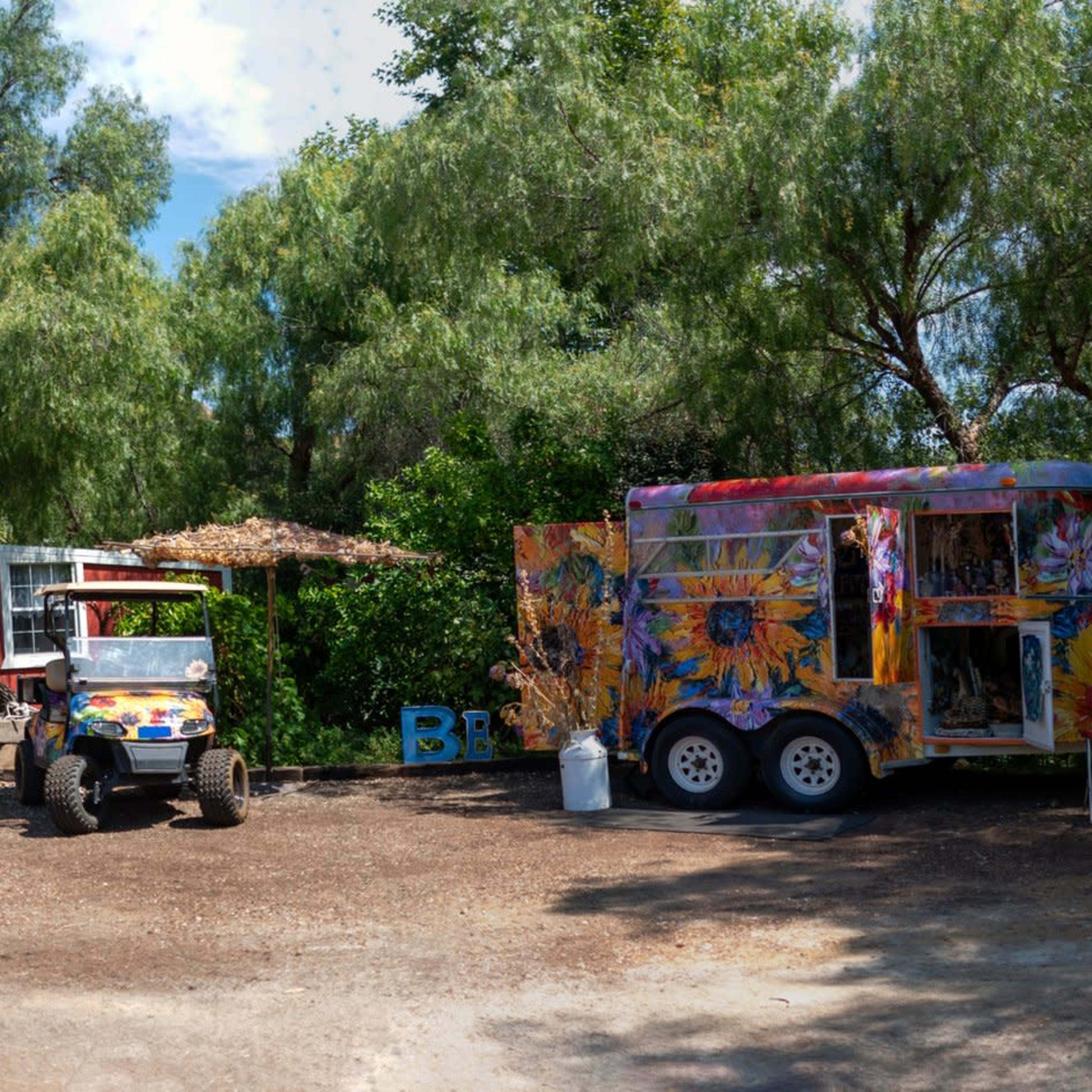 A colorful outdoor scene featuring a vibrant food truck, a white horse sculpture, a parked golf cart, and a red barn surrounded by lush greenery and palm trees.