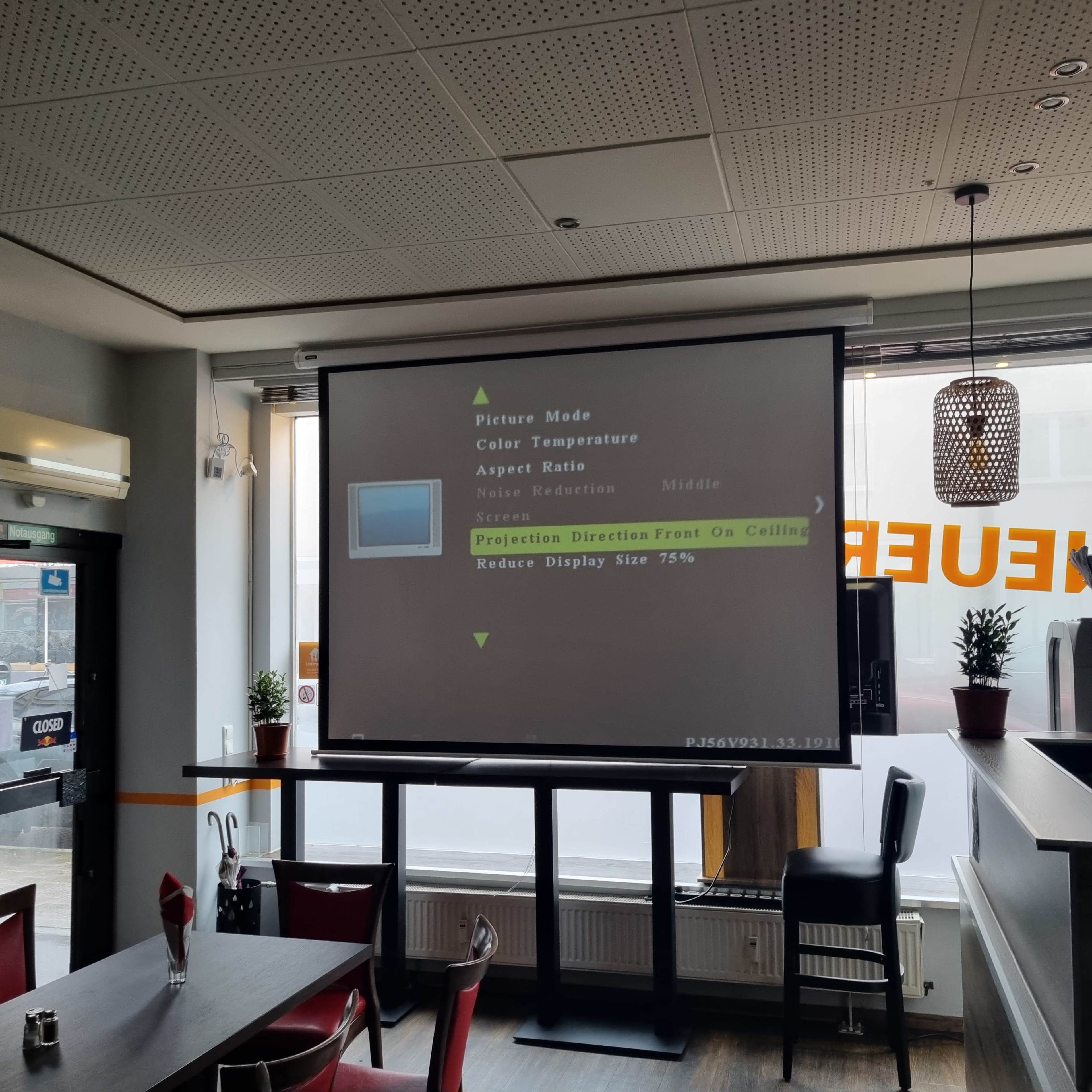 A large projector screen displays settings menus in a cafe setting.