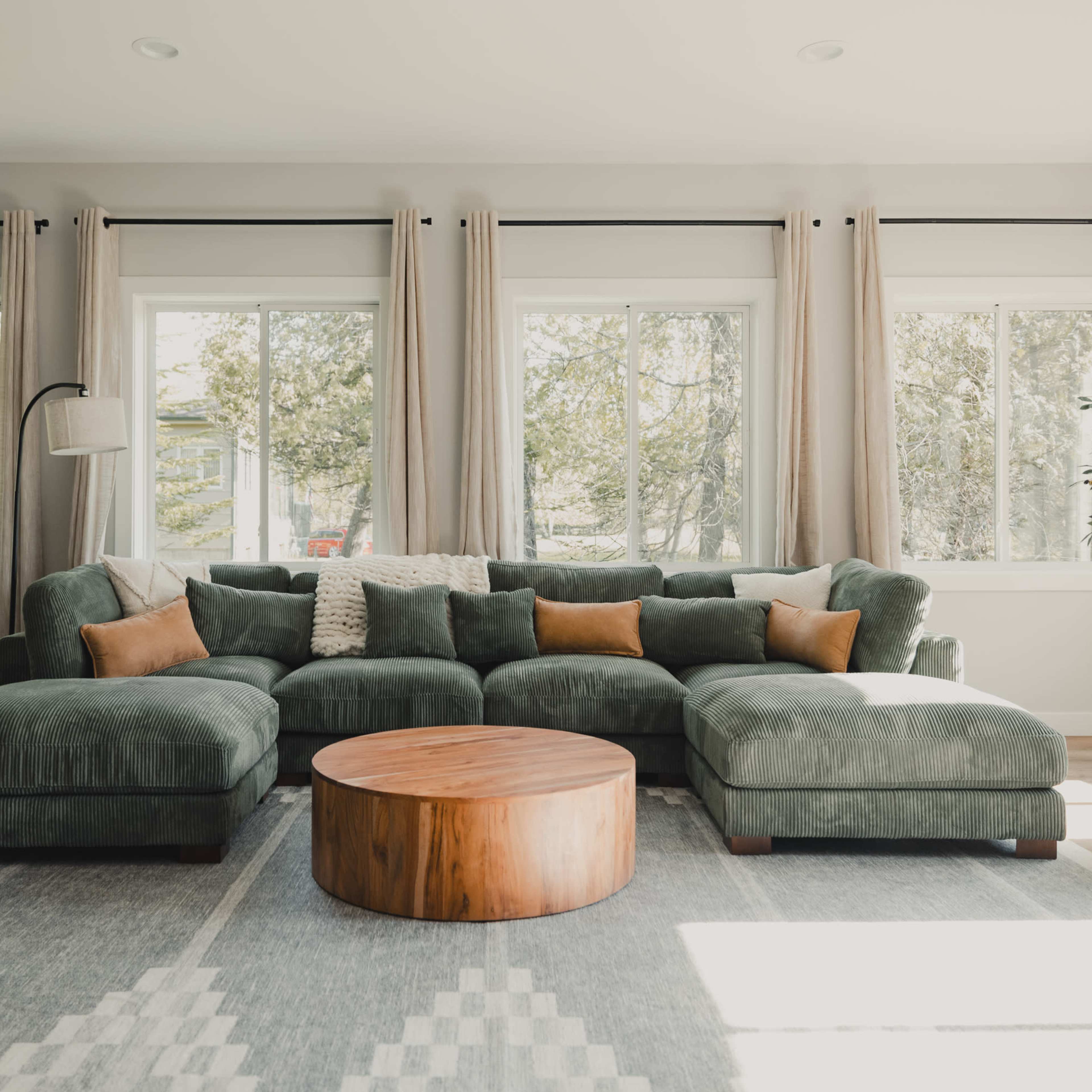 A green sectional sofa with brown accent pillows is placed in a brightly lit living room with large windows and a round wooden coffee table.