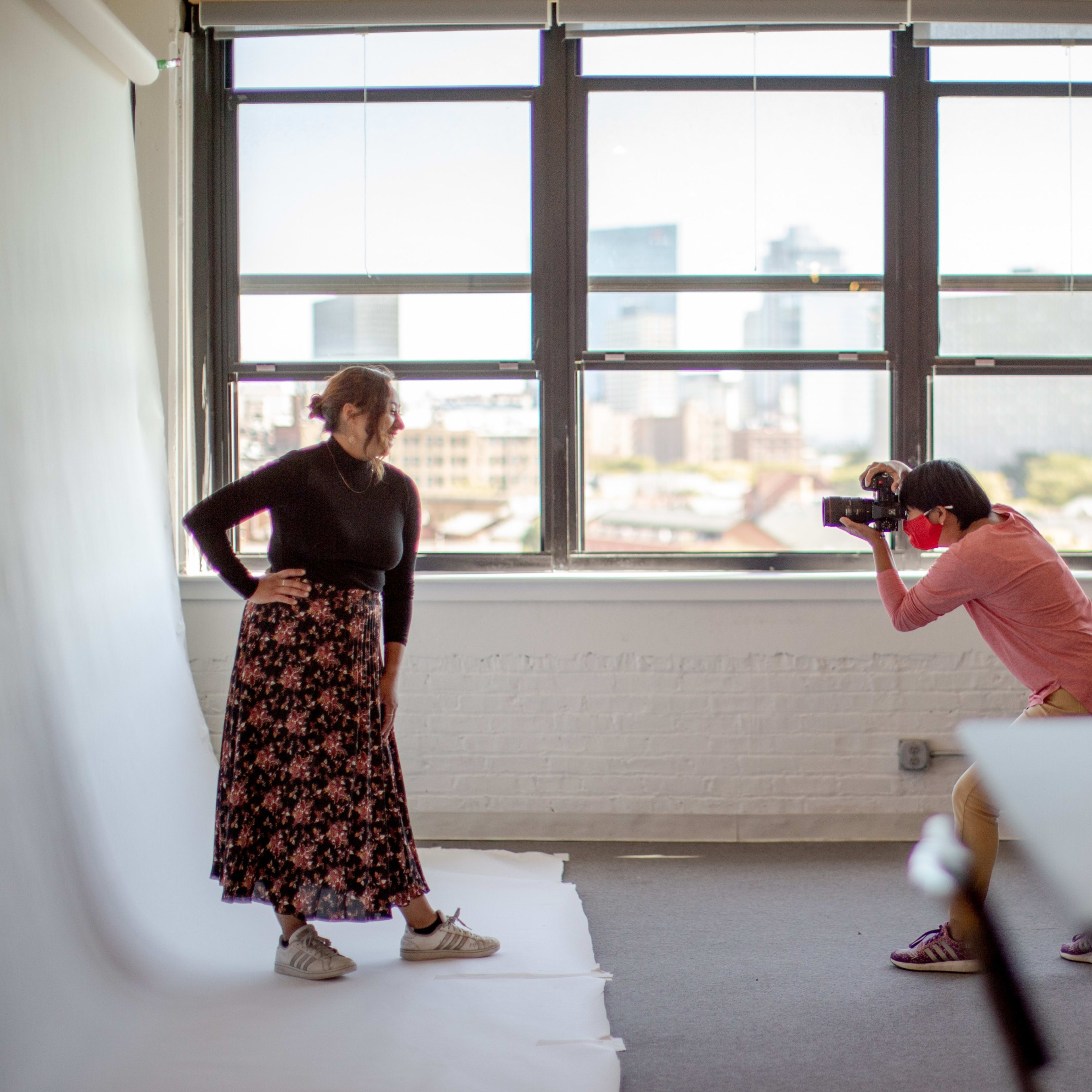 A photographer captures a model posing with a floral skirt in a sunlit studio with large windows and a city skyline view.