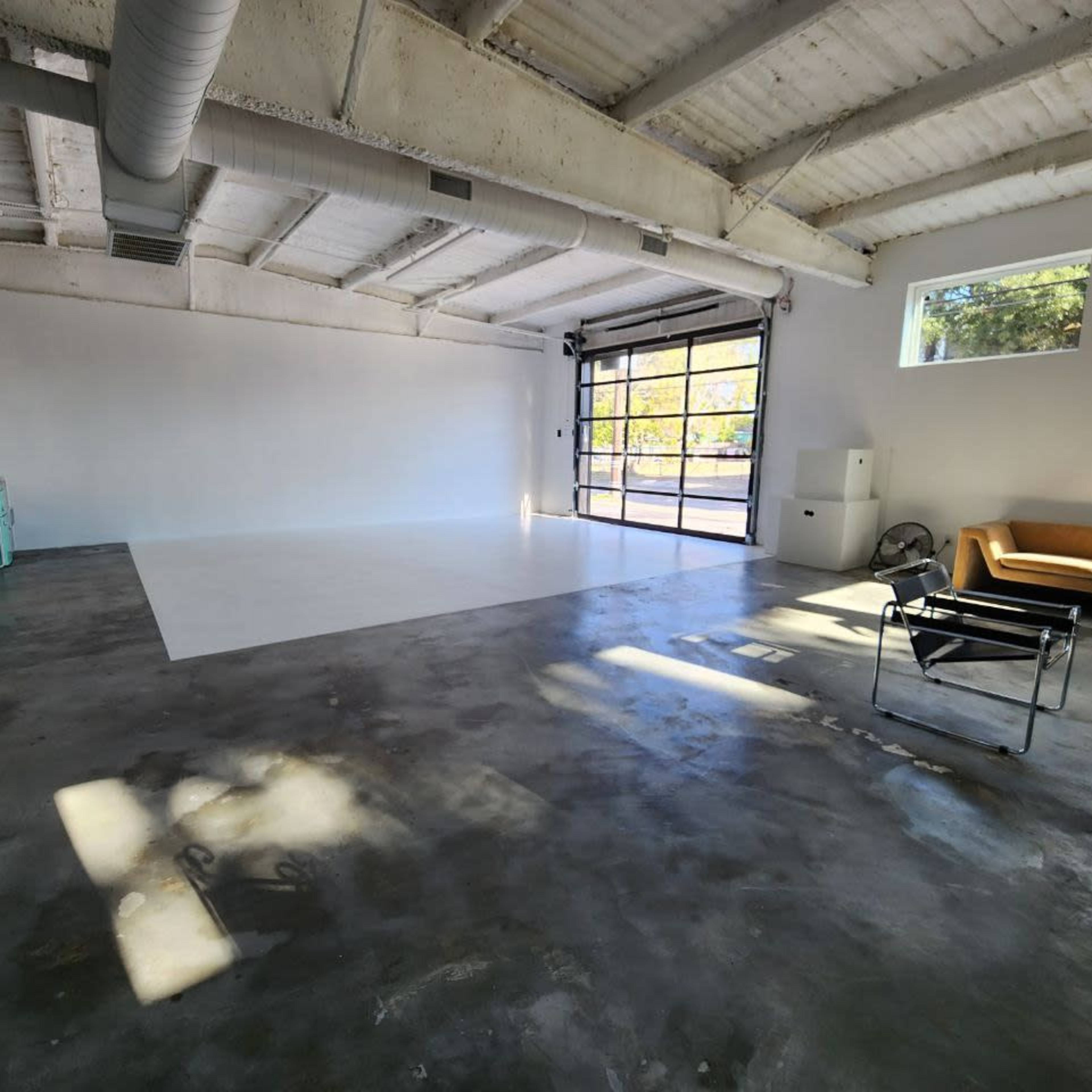 A spacious, empty studio with a polished concrete floor, large windows, and minimal furniture, including a couch and a chair.