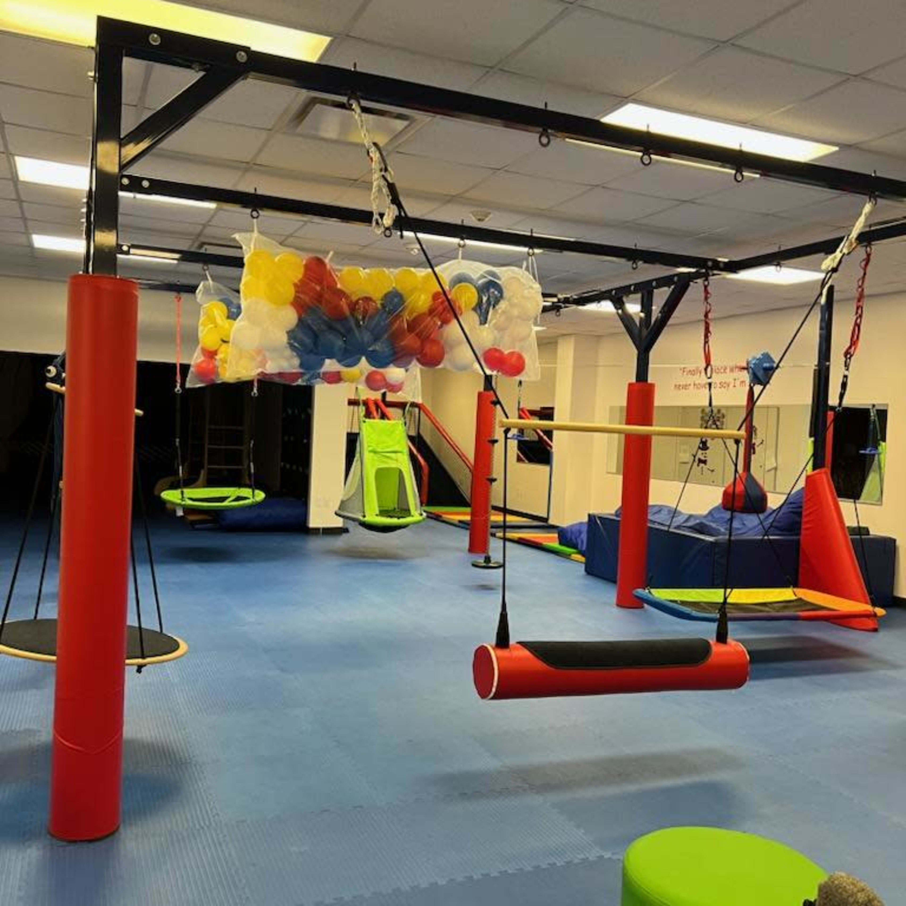 An indoor play area equipped with various colorful equipment, including swings and climbing structures, set against a rubber mat flooring.