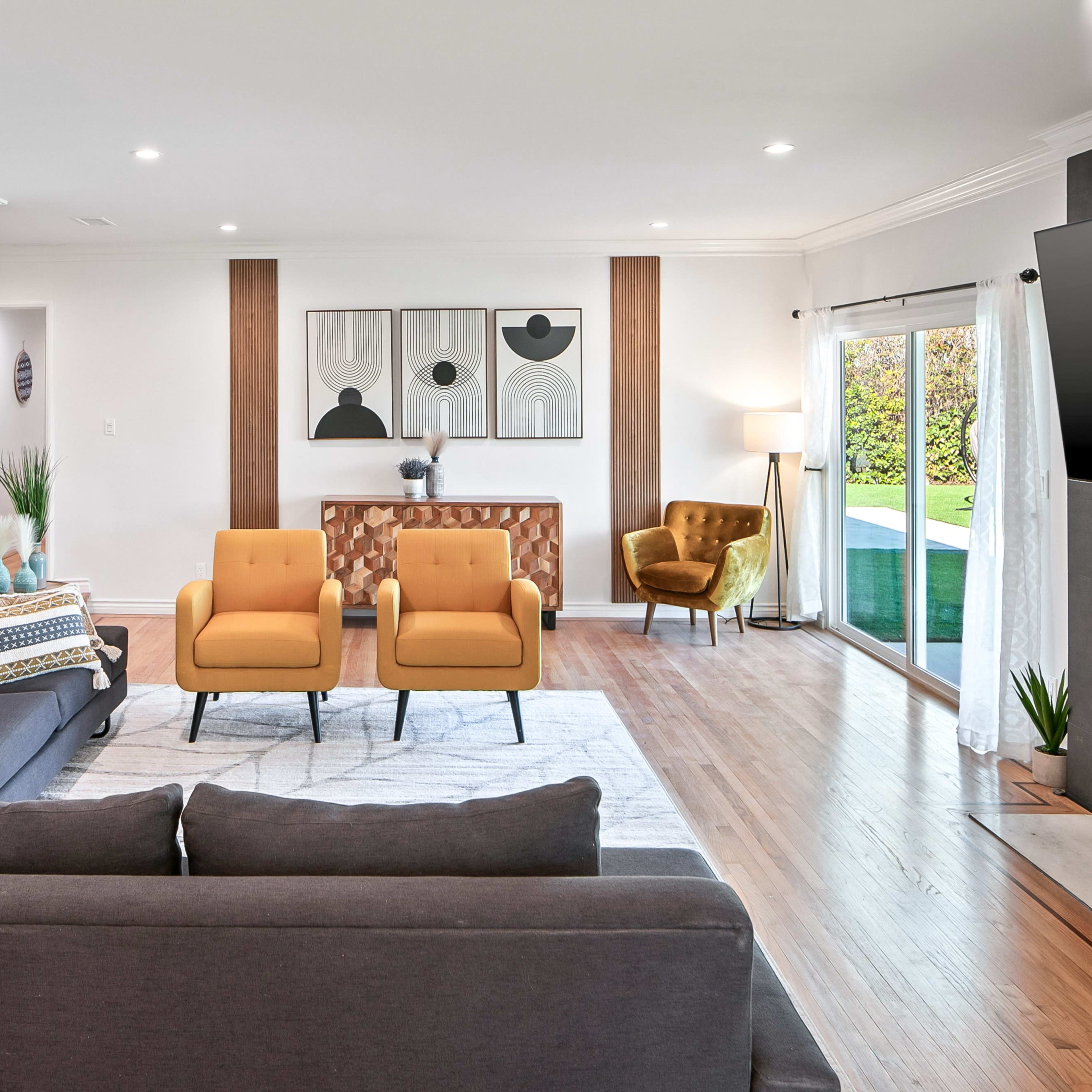 The living room features a gray sectional sofa, two yellow armchairs, and a console table against the wall, with large windows providing a view of the garden.