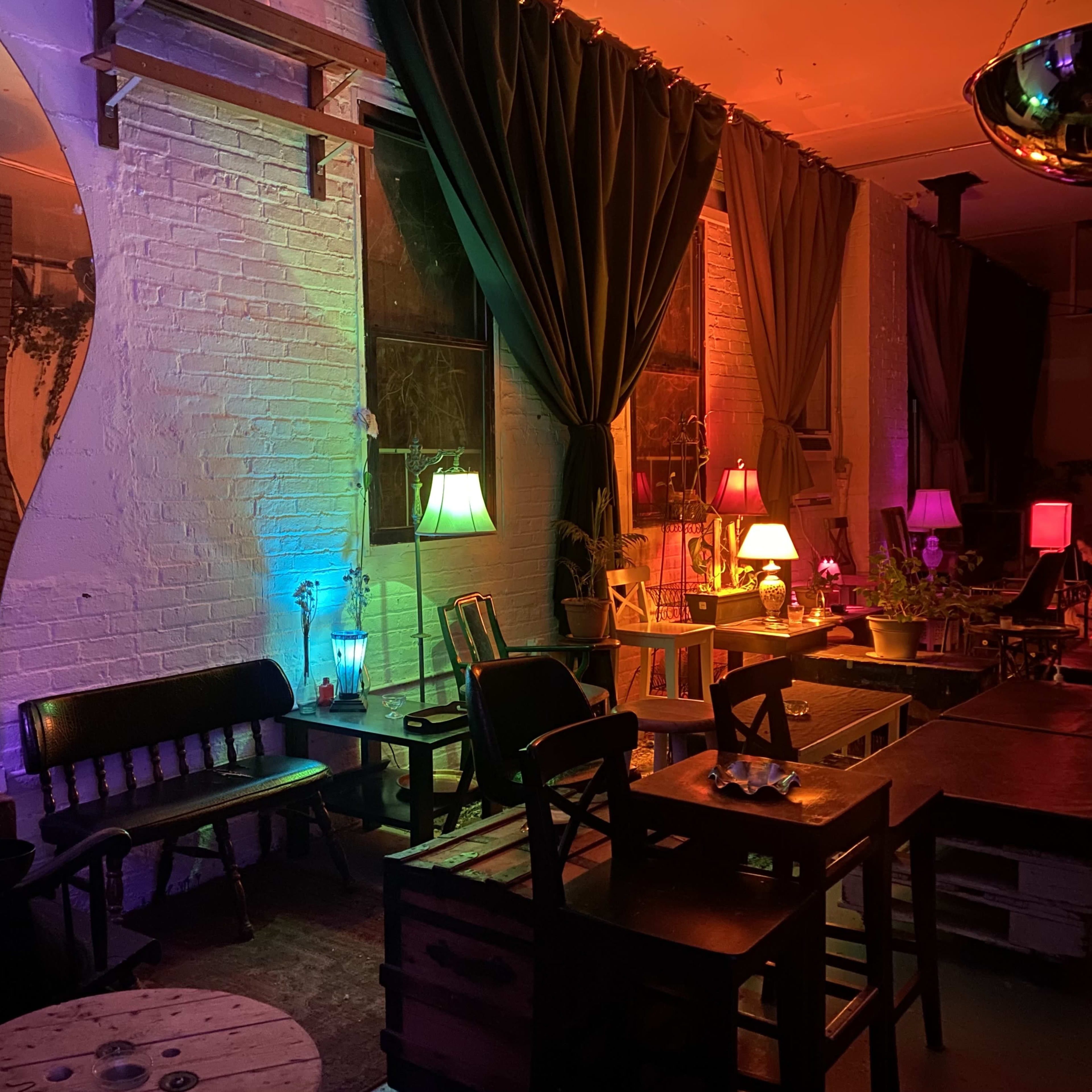 A dimly lit lounge area featuring colorful lighting, wooden furniture, and various lamps scattered throughout.