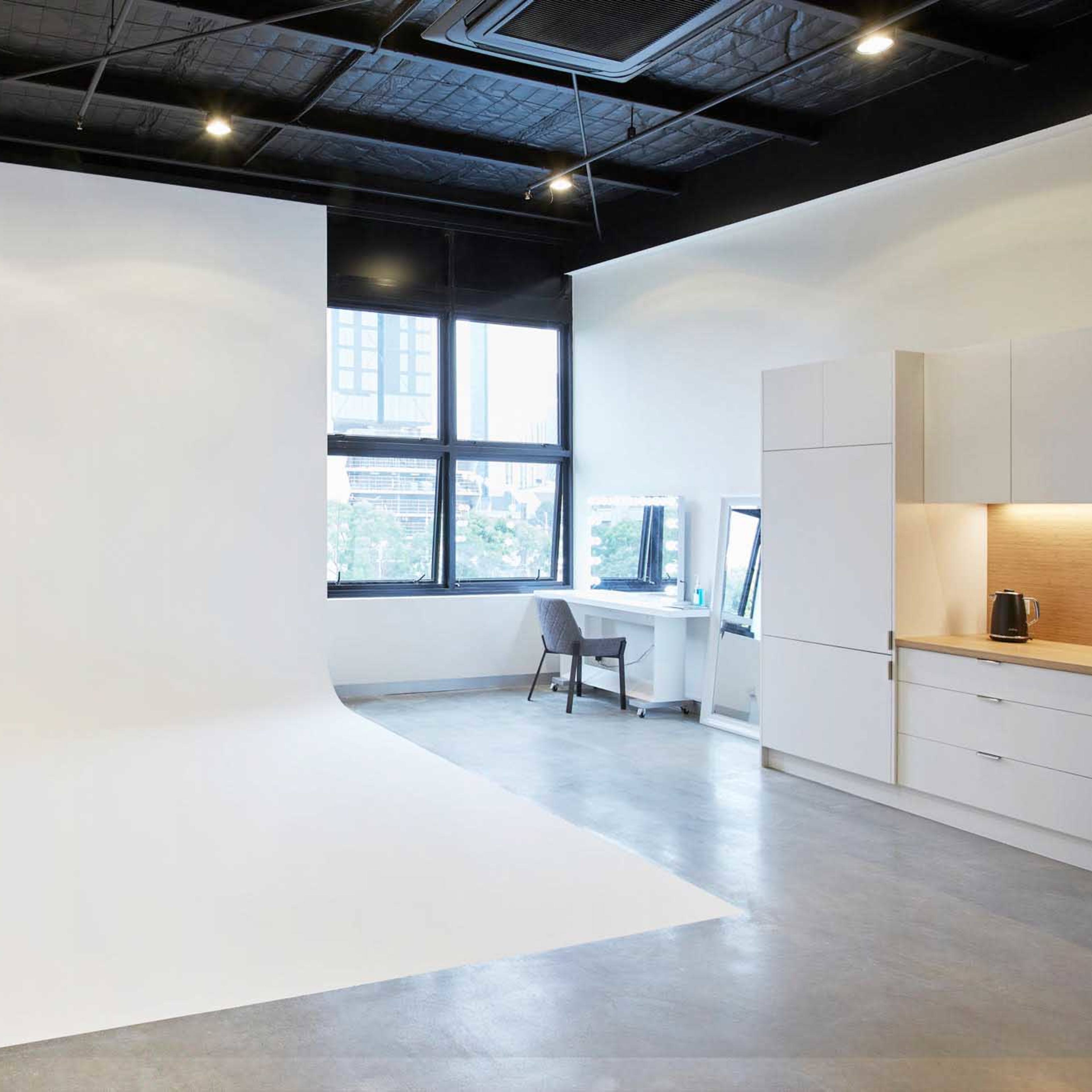 A minimalistic photography studio with a large white backdrop, a desk, and a kitchenette in a brightly lit room.