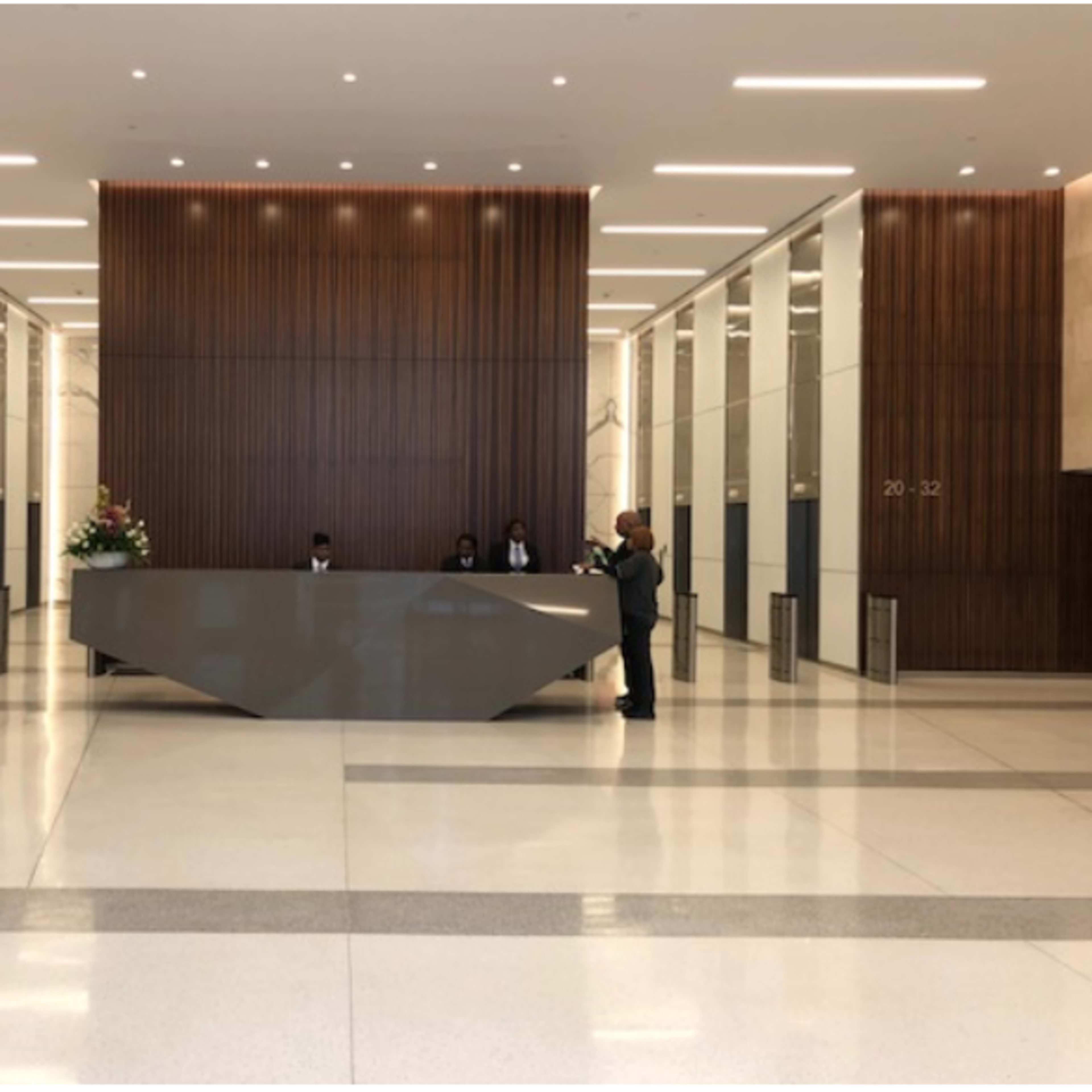 A modern lobby with a reception desk and four attendants standing in front of it, surrounded by polished floors and panelled wooden walls.