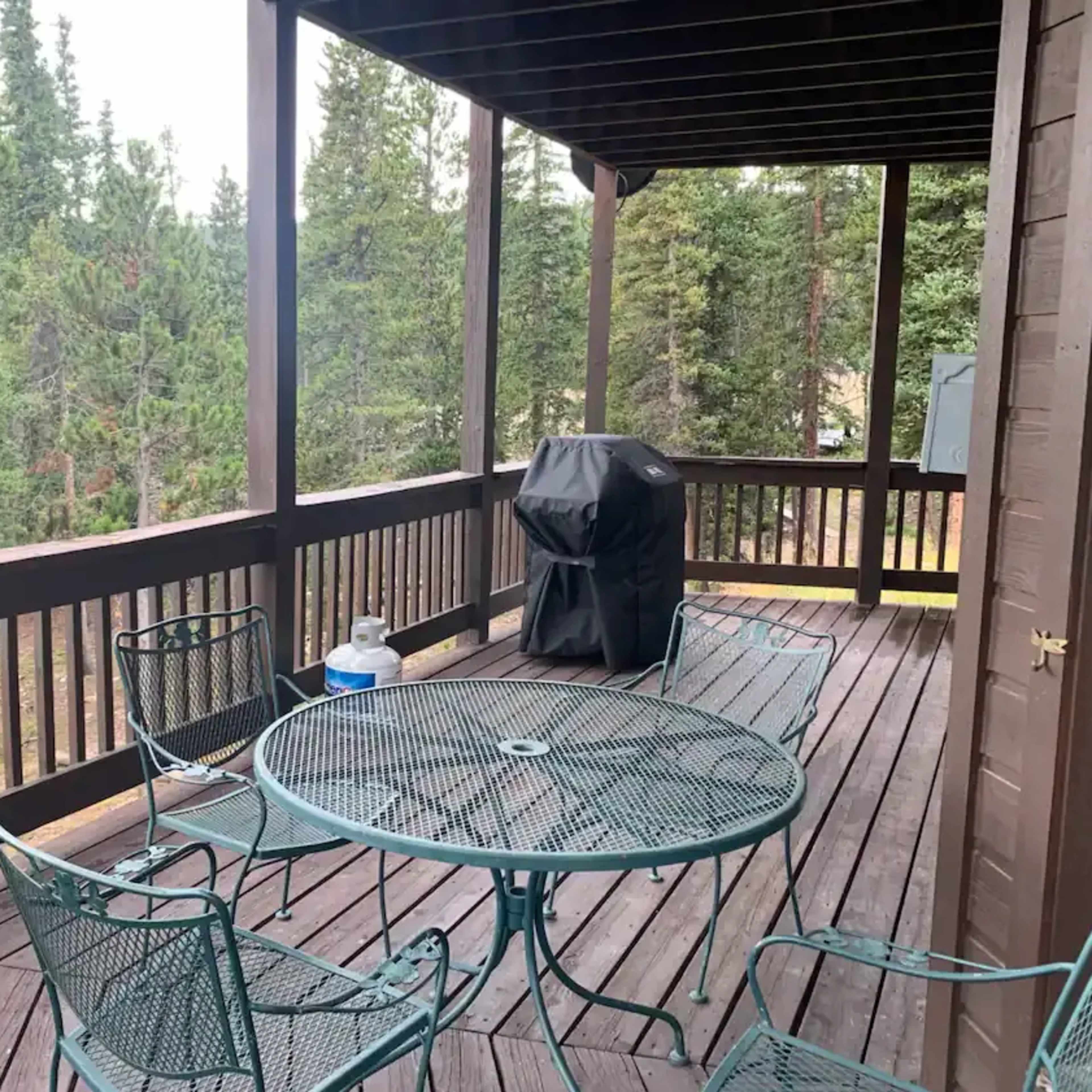 A wooden deck with a circular metal table and four chairs overlooks a forested area, featuring a grill in the corner.