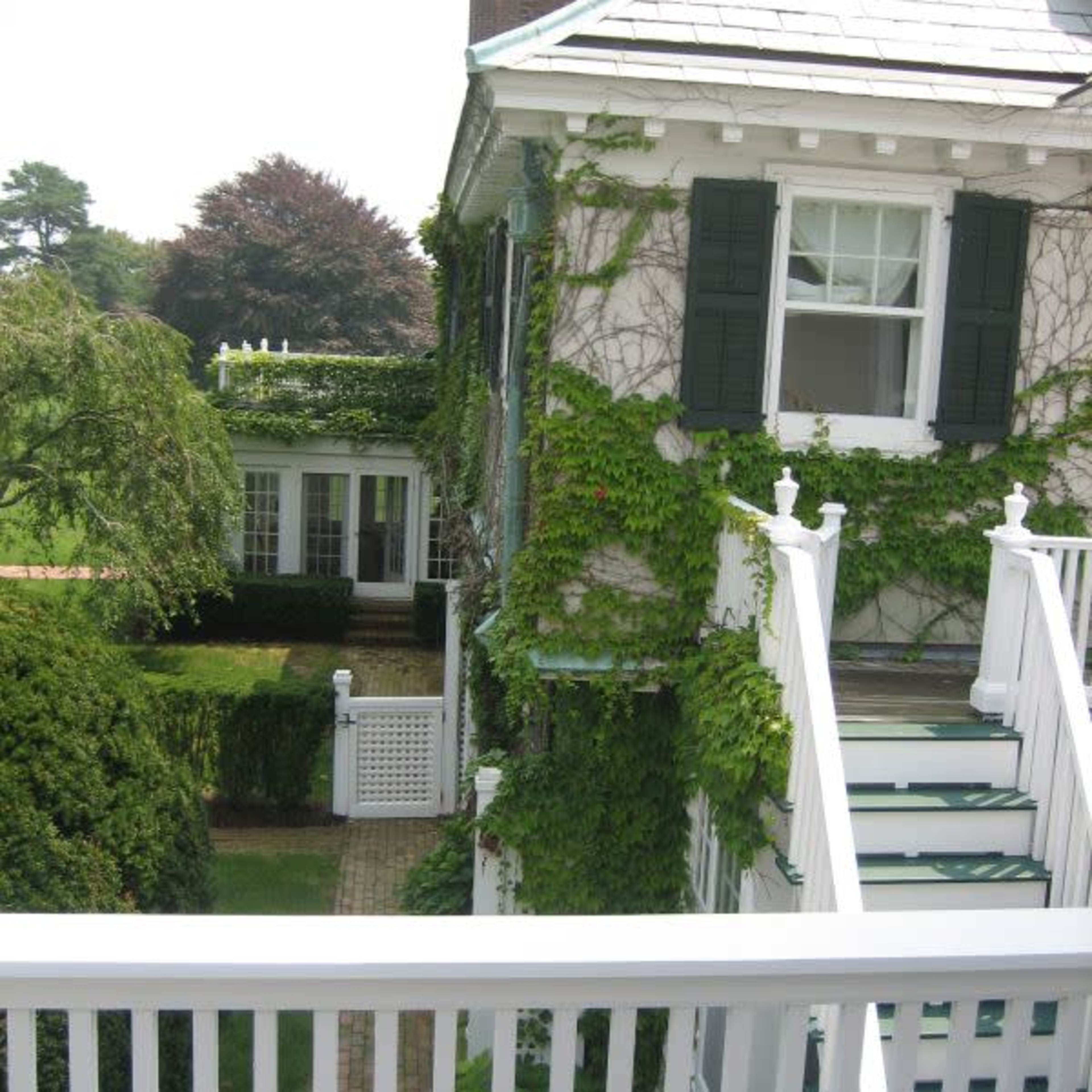 A two-story house covered in green ivy, with white railings and a glimpse of a landscaped yard in the background.