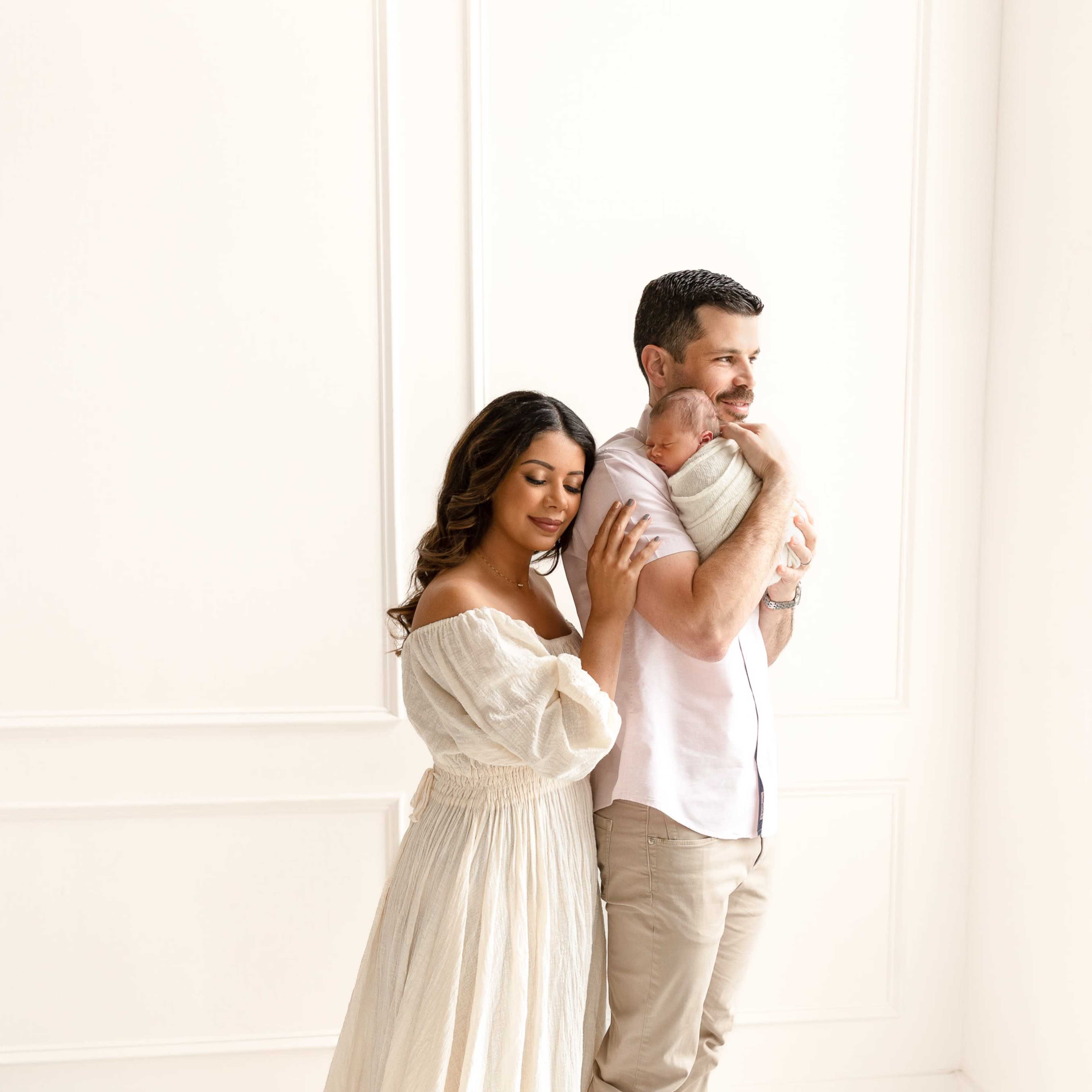 A woman in a flowing dress stands beside a man who is holding a baby, both looking towards the camera in a softly lit room.