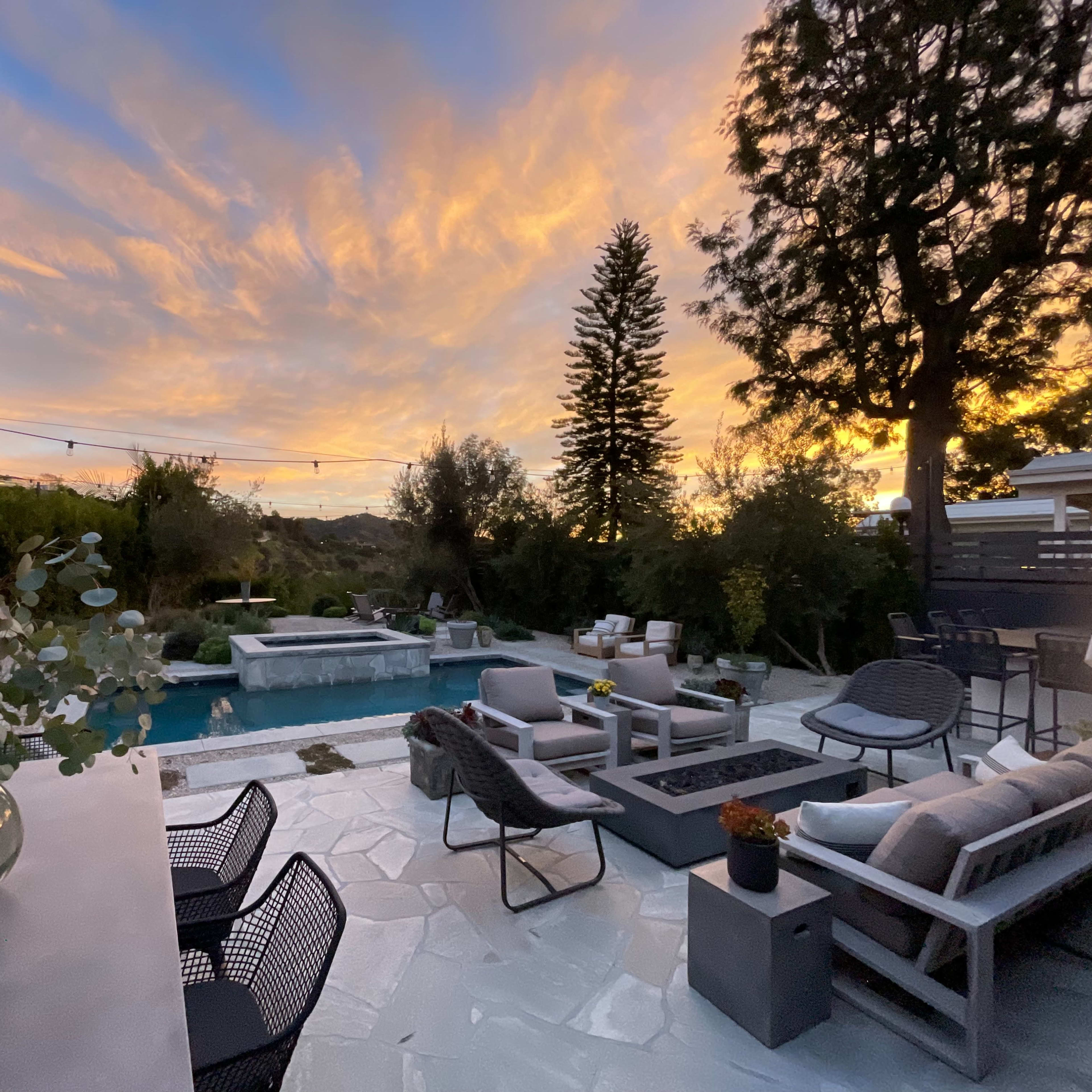 A backyard patio with a pool, seating areas, and a beautiful sunset sky filled with clouds.