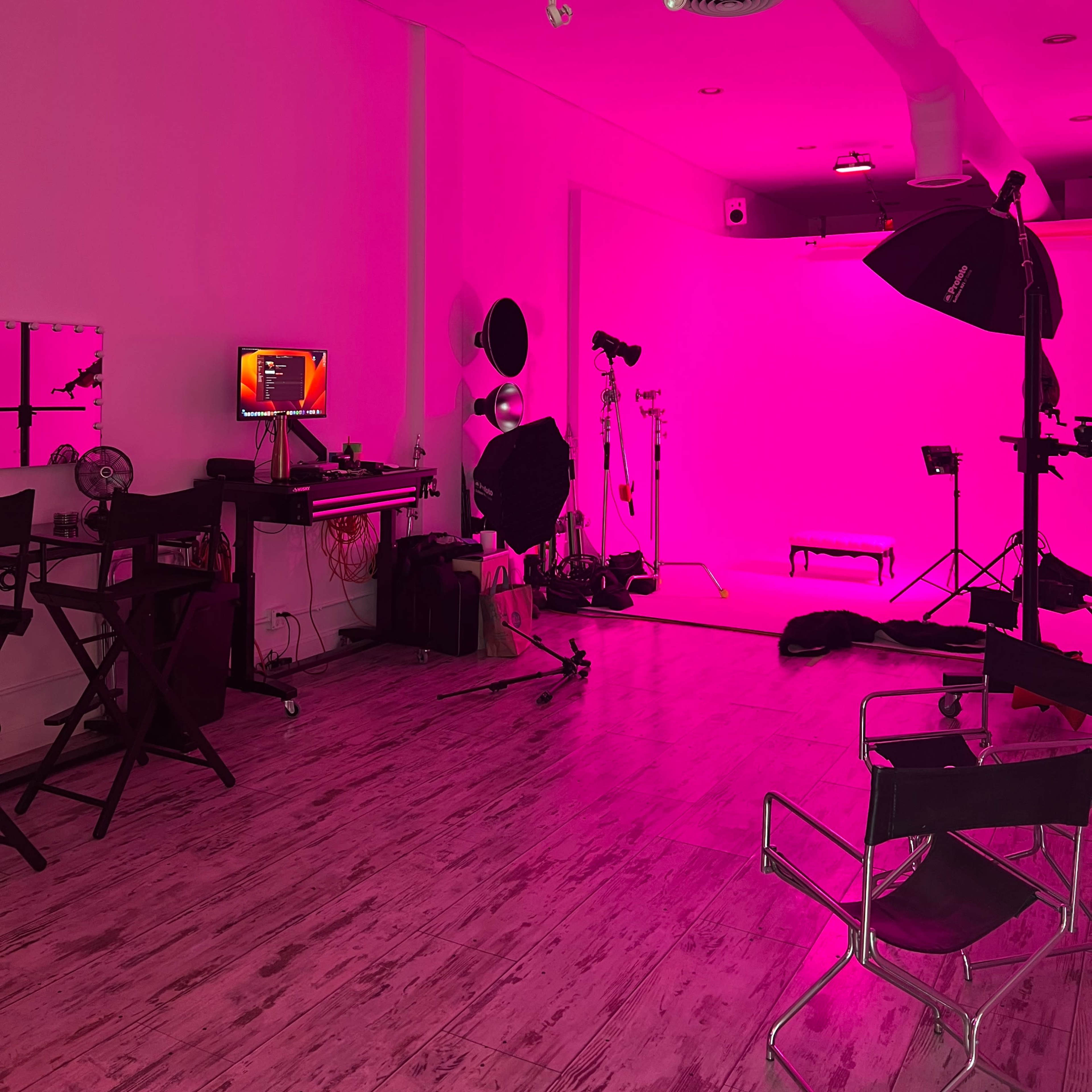 A photography studio is illuminated with pink lighting, featuring various equipment including softboxes, a backdrop, and a computer setup.