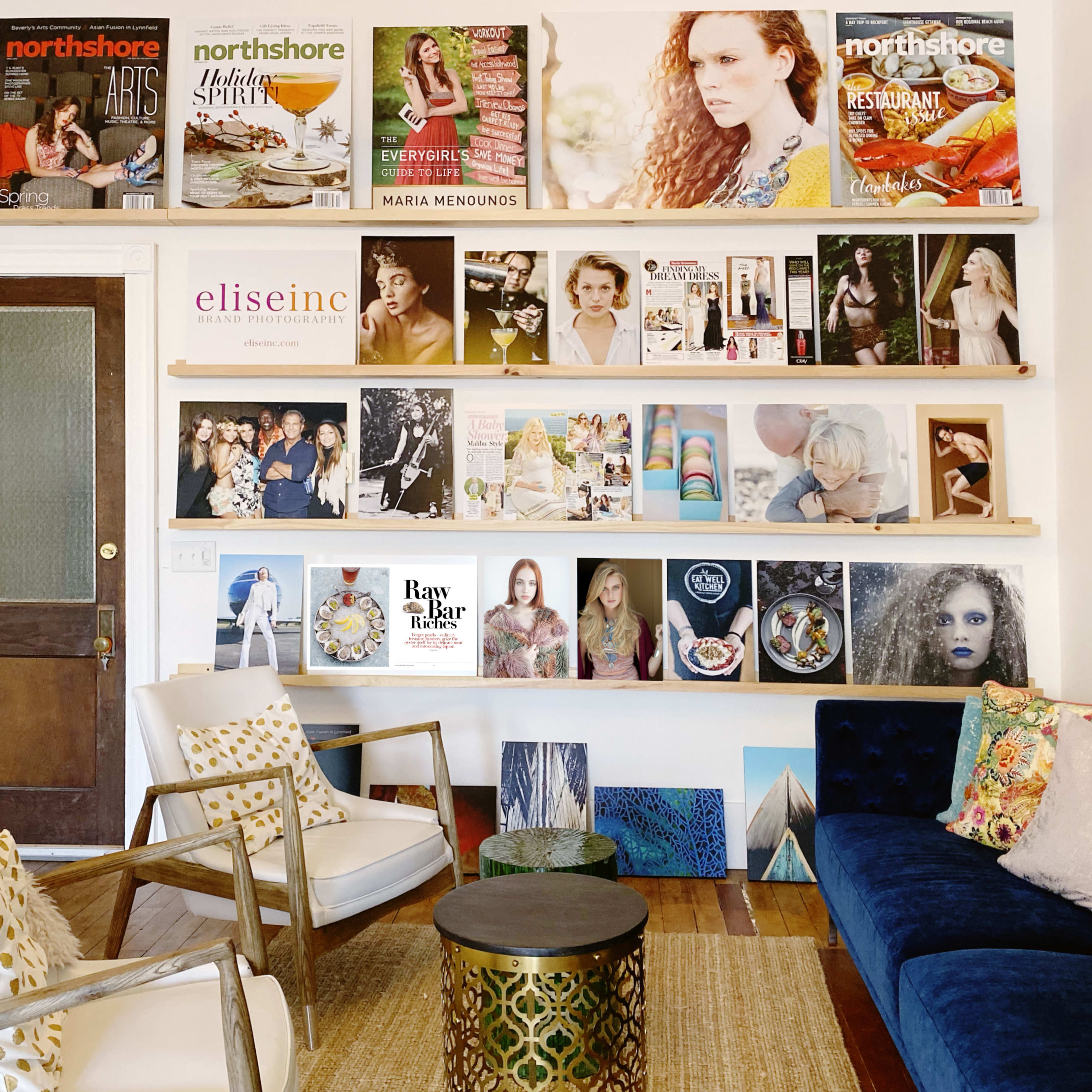 A stylish interior with a wall displaying magazines and artwork, complemented by two chairs and a sofa adorned with colorful cushions.