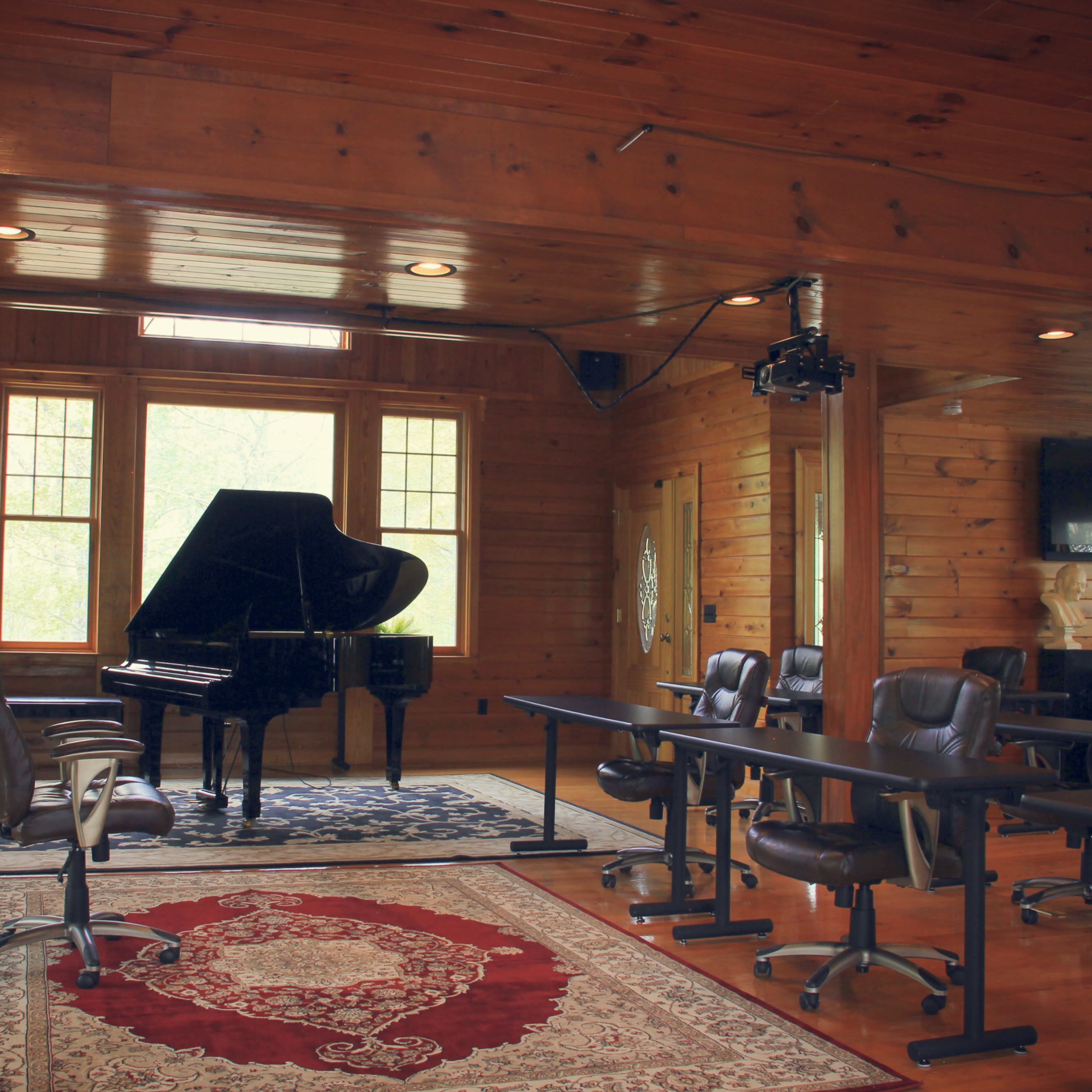 A wooden room with a grand piano, several chairs, and tables arranged for a meeting or class, illuminated by natural light from large windows.