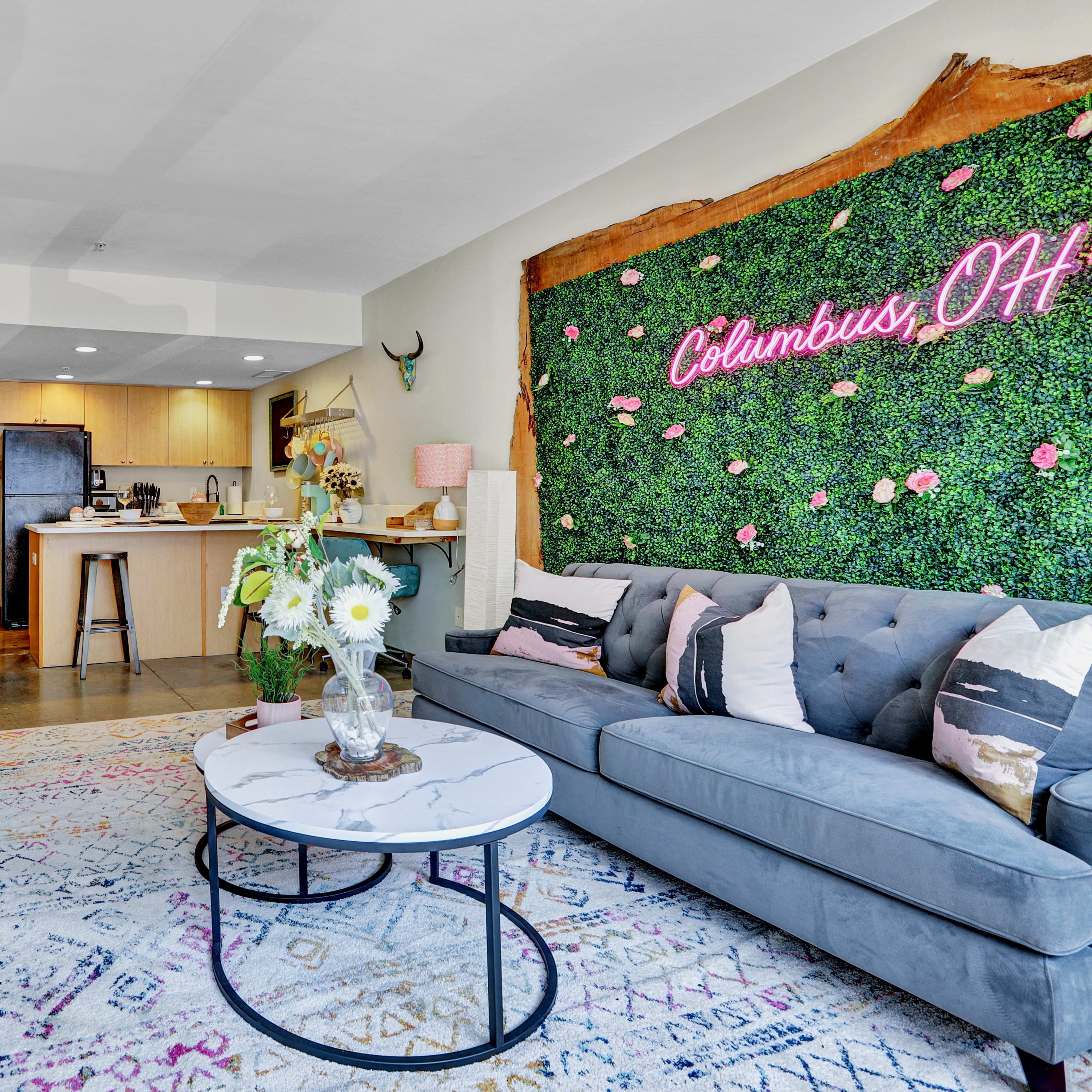 A living room with a gray sofa, a round coffee table, a decorative green wall featuring the text "Columbus, OH," and an open kitchen in the background.