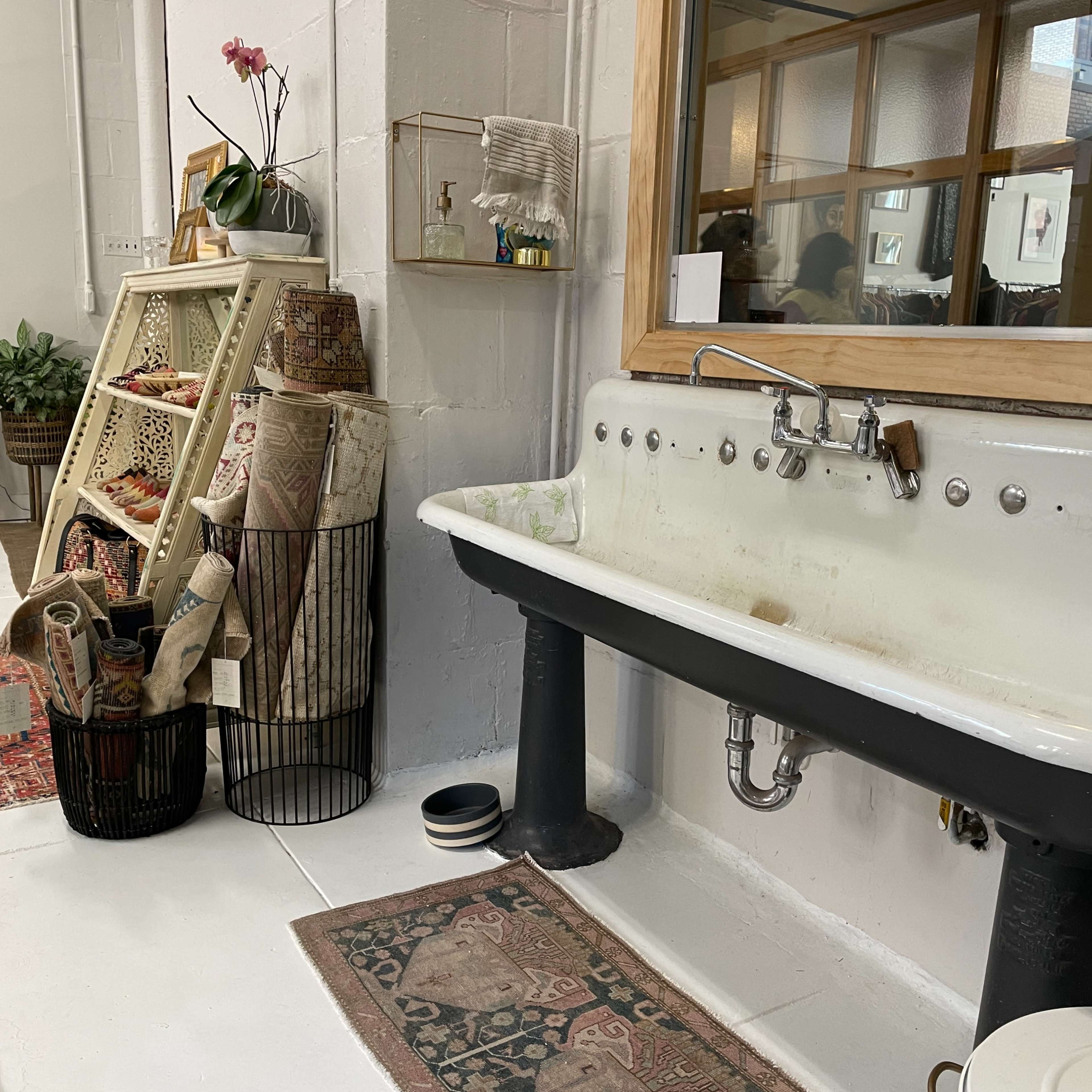 A bright, minimalistic room featuring a vintage sink, an assortment of textiles displayed on a rack, and a few decorative elements on the walls.