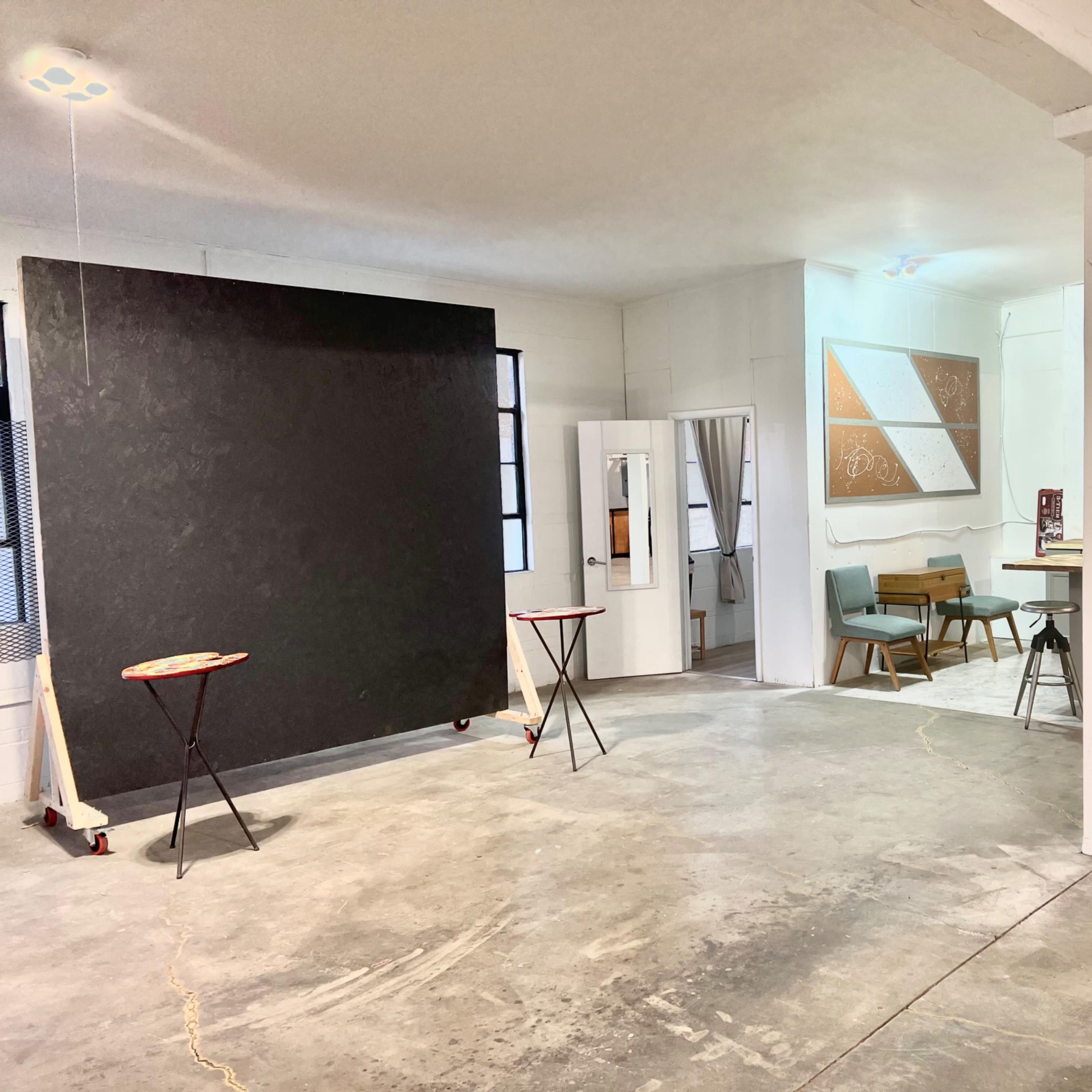 An open studio space featuring a large black wall, two small tables, a kitchenette, and seating in a well-lit area.