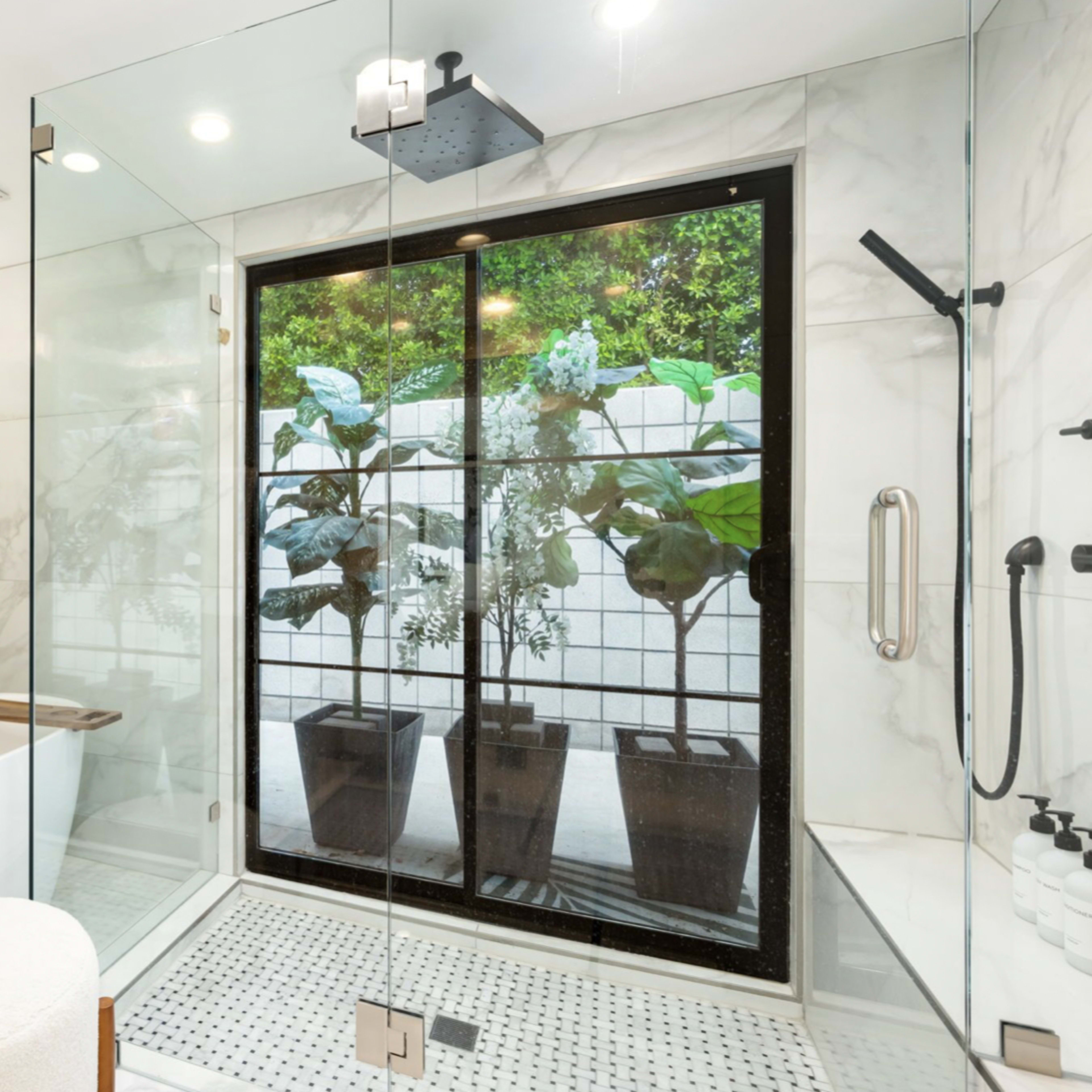 A modern bathroom featuring a glass shower enclosure, a freestanding bathtub, and large windows with potted plants outside.