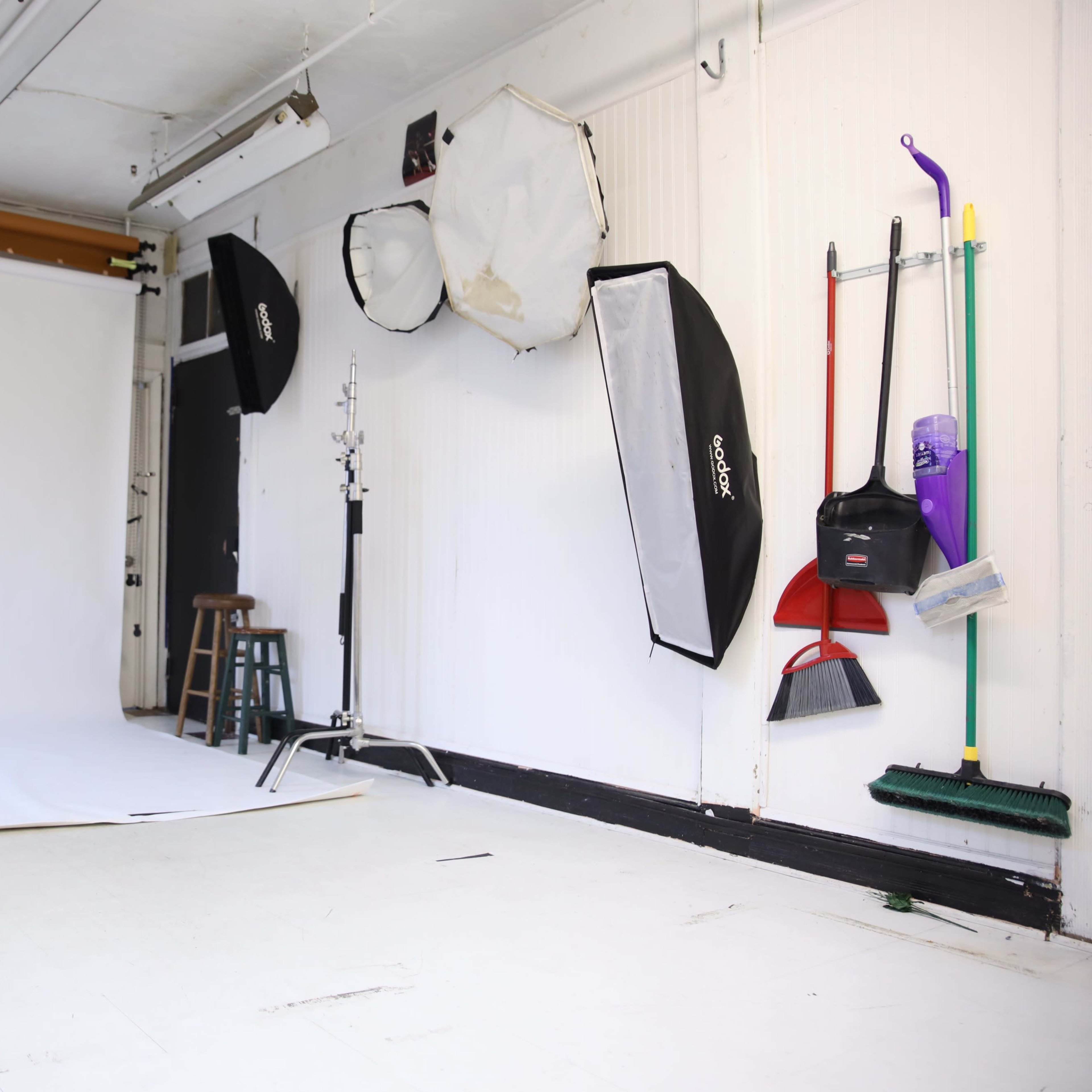 A photography studio with a white backdrop, various light modifiers, and cleaning tools hanging on the wall.