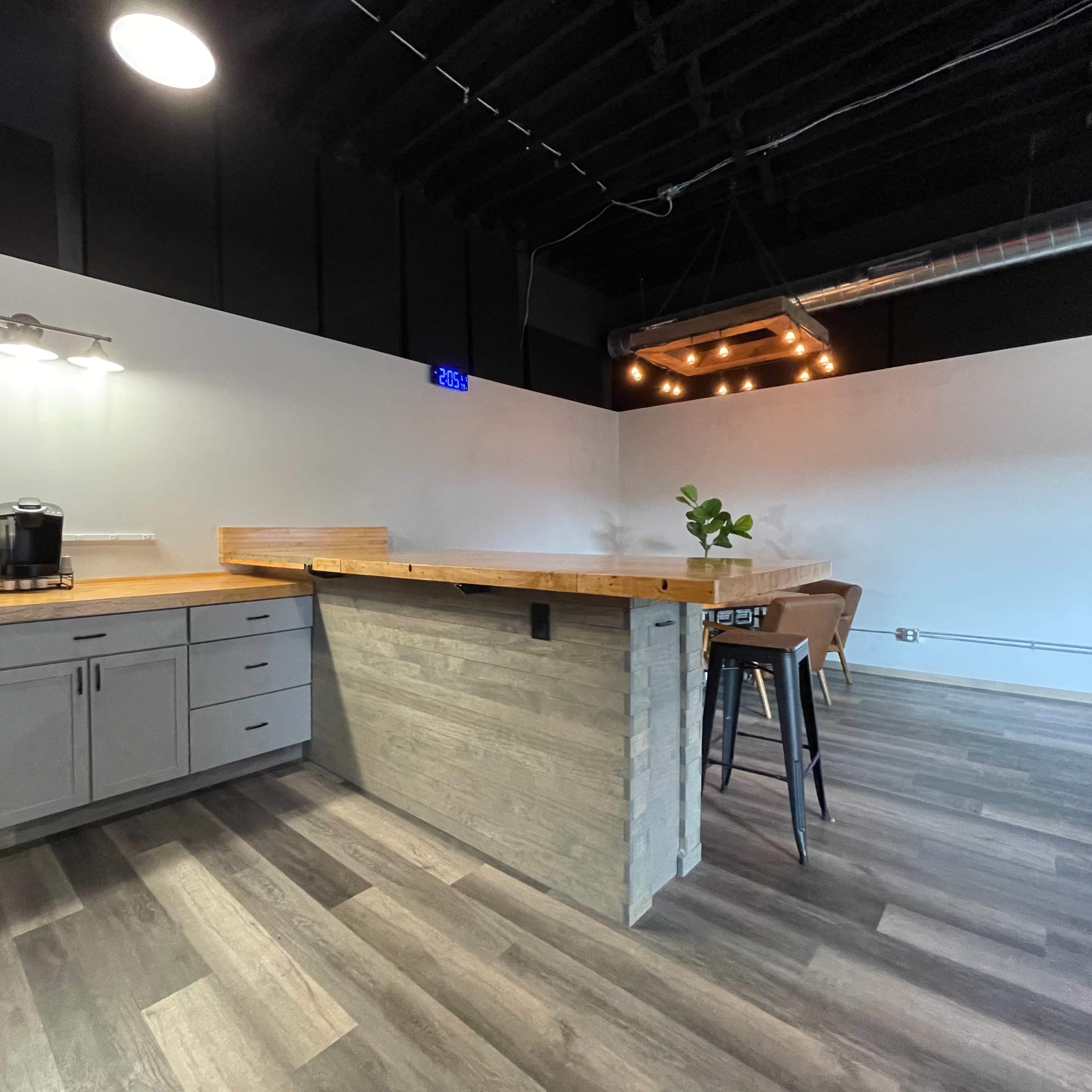 A modern kitchen space with a wooden bar counter, cabinetry, and a coffee maker against a minimalist backdrop.