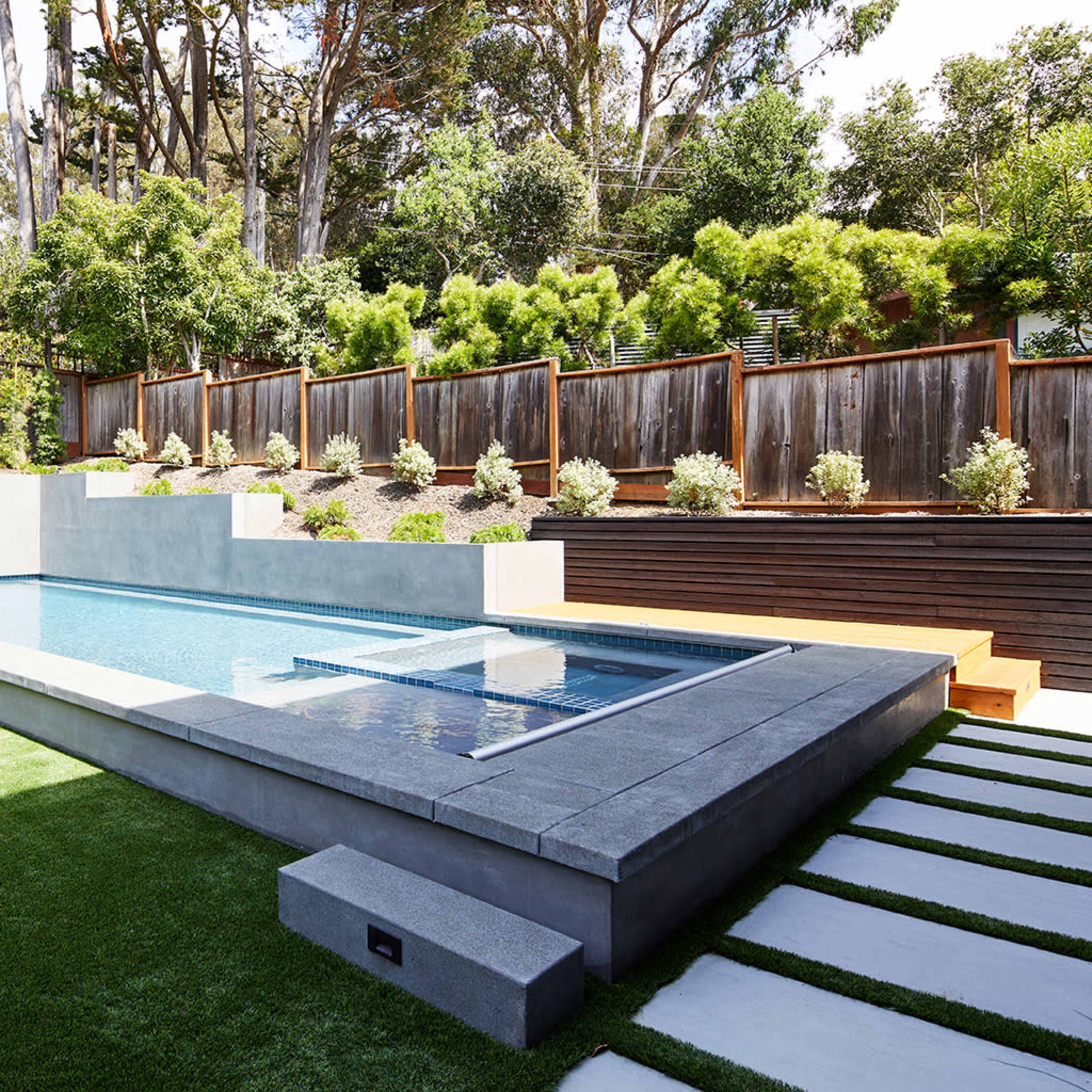 A modern swimming pool surrounded by landscaped greenery and a wooden fence, with patio seating and stone walkways.