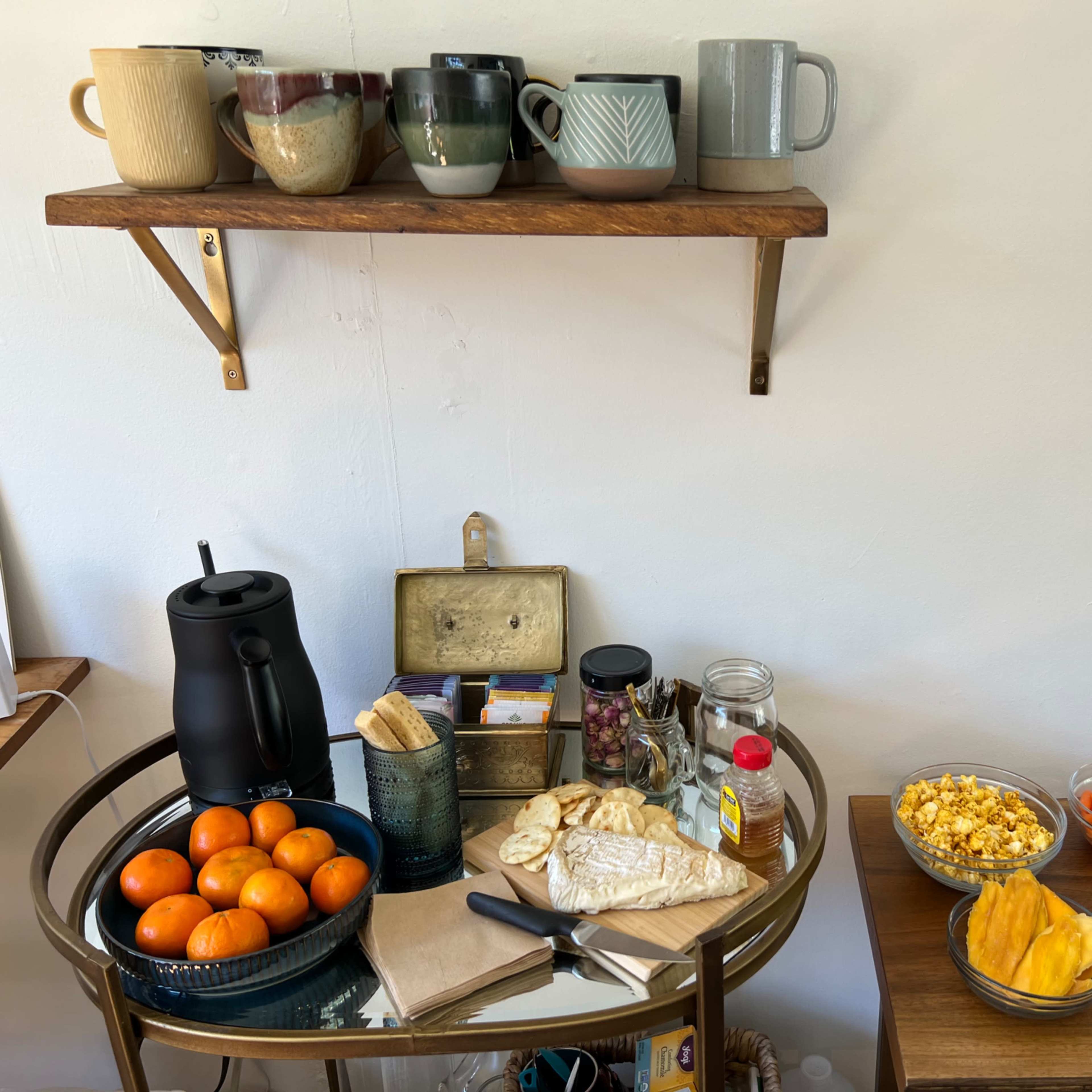 A wooden shelf holds several mugs above a table laden with snacks, fruit, and beverages, including tea, popcorn, and fresh oranges.