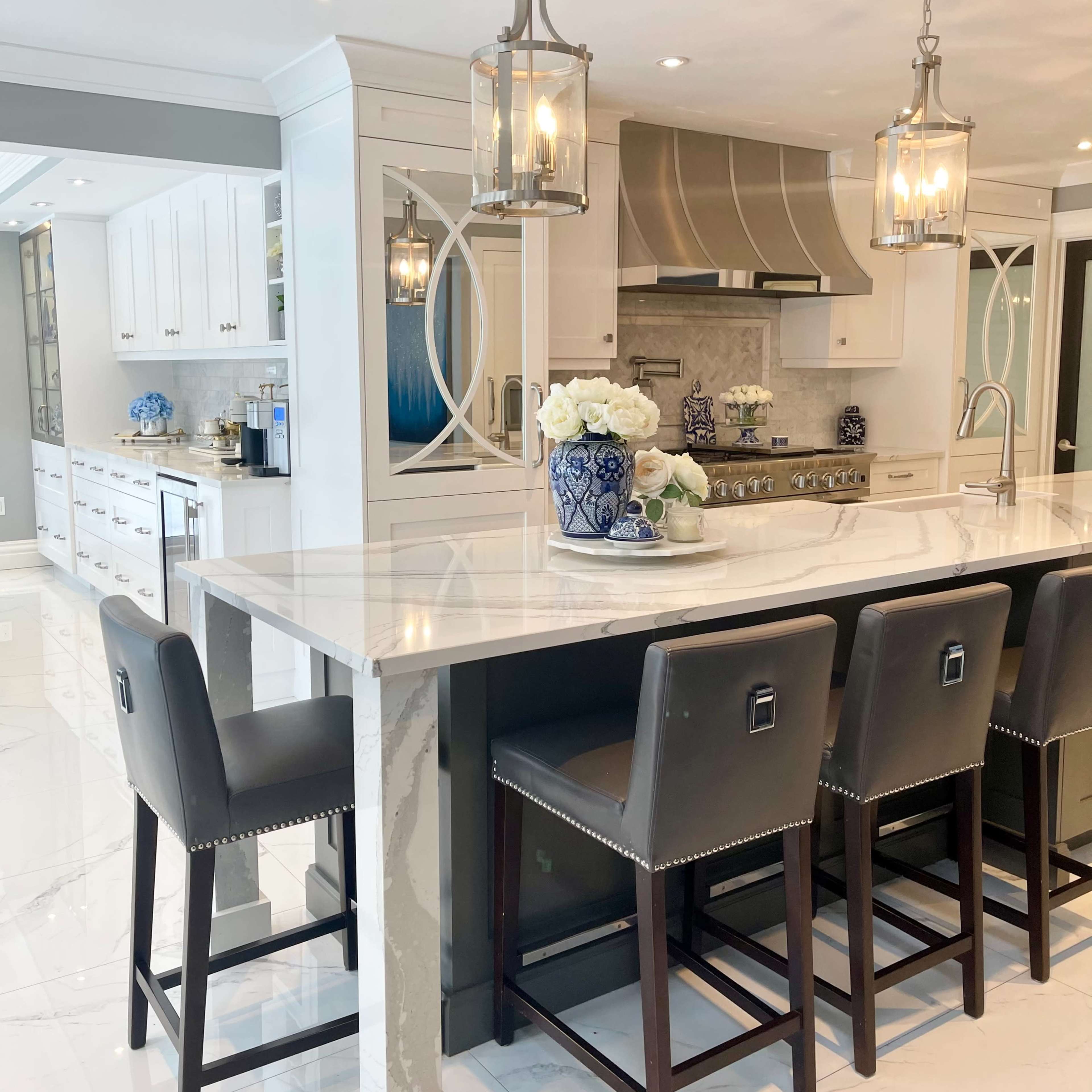 A modern kitchen features a large marble island with bar stools, silver pendant lights, and white cabinetry.