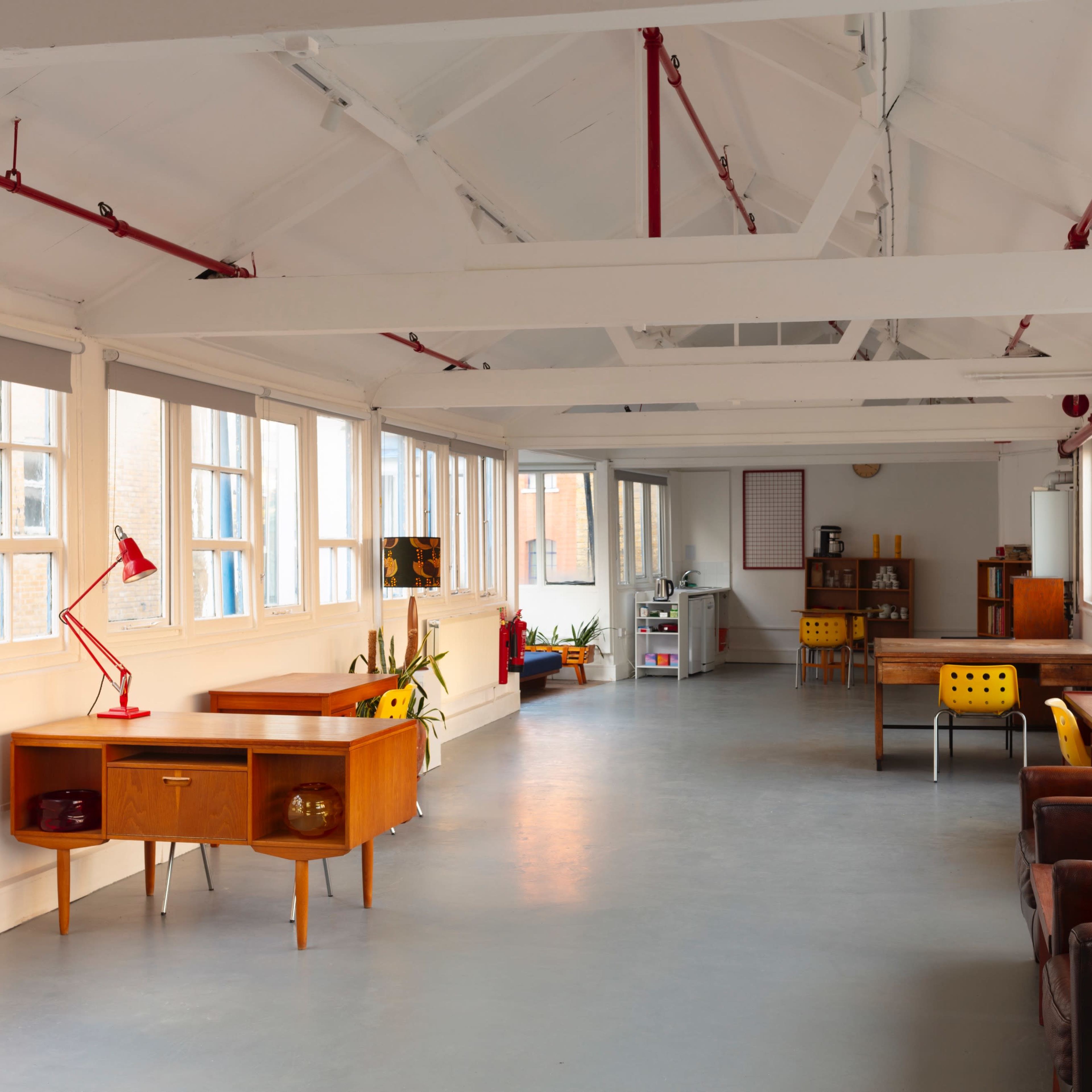 The interior of a bright, open space features wooden furniture, including desks and chairs, with large windows allowing natural light to enter.