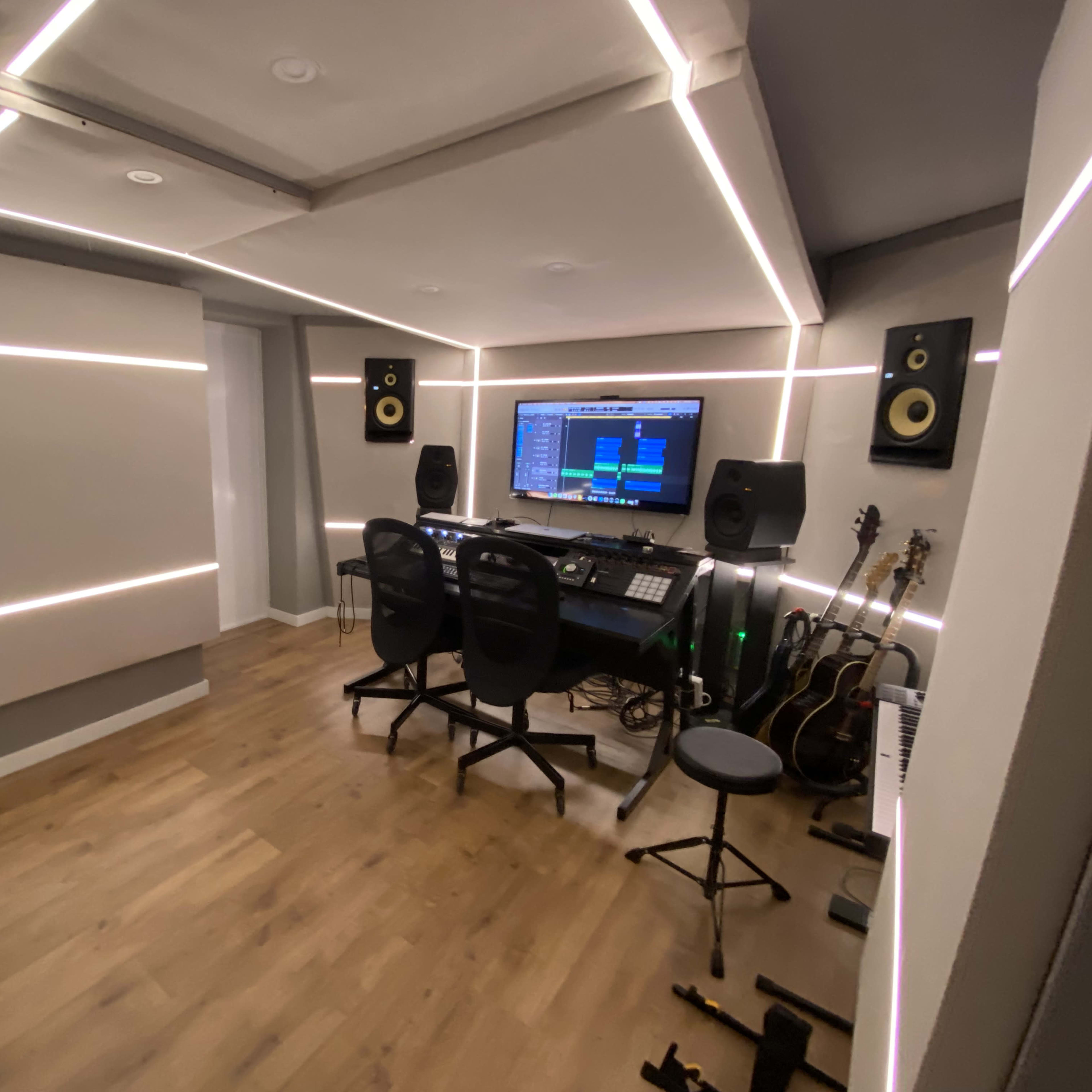 A music studio with a digital audio workstation setup, speakers, a seating area, and several guitars leaning against the wall.