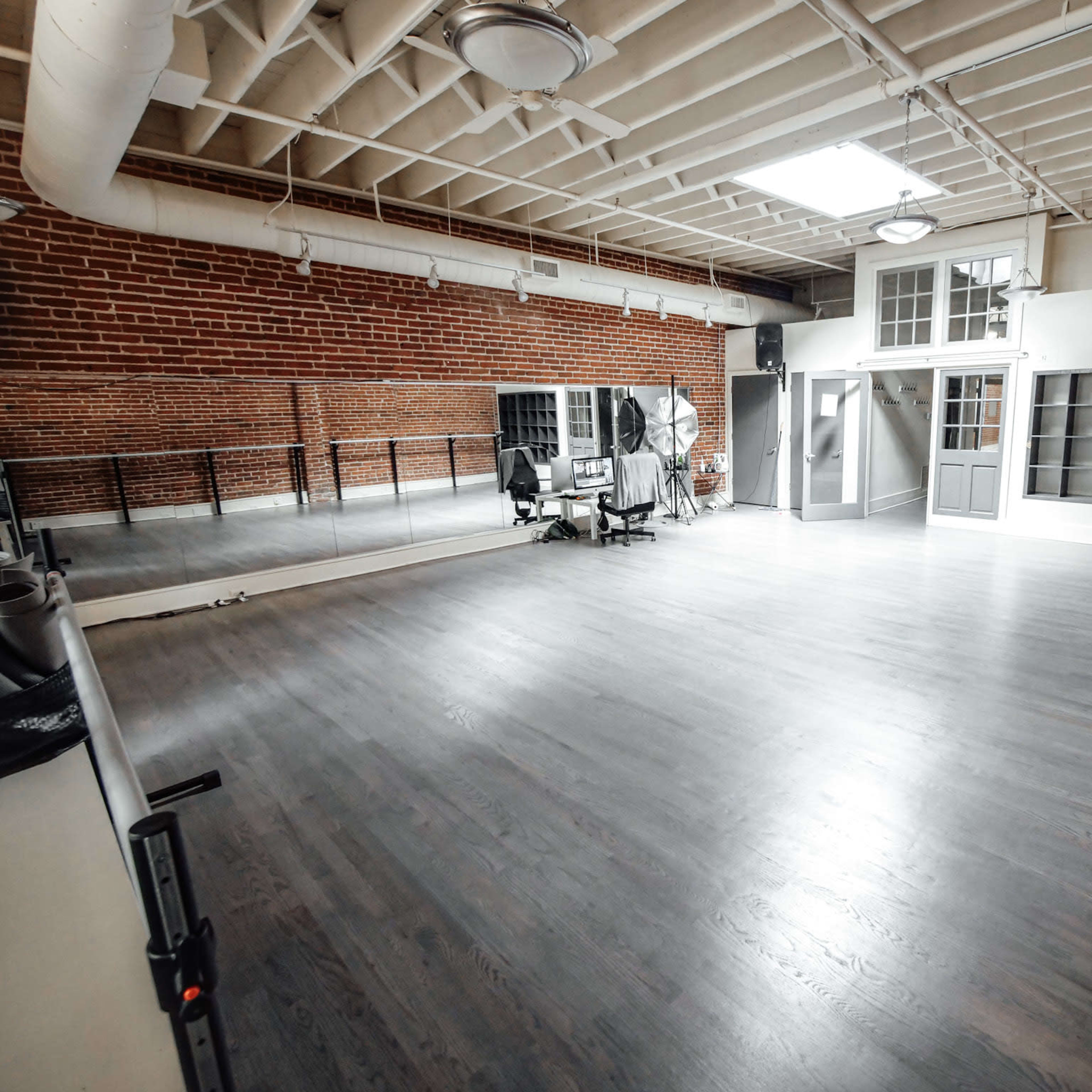A spacious, well-lit dance studio with wooden floors, exposed brick walls, and a large mirror along one side.