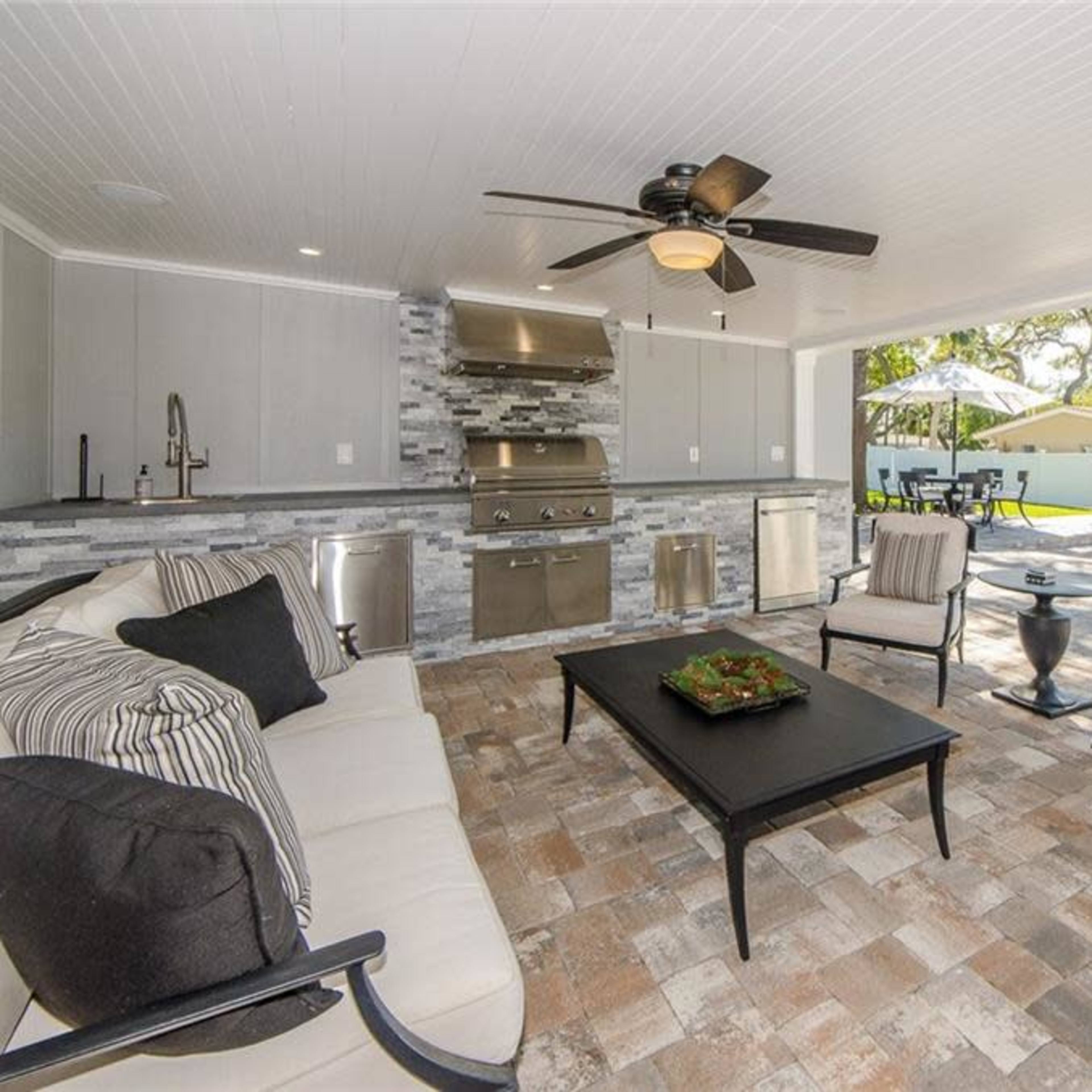 A covered outdoor living space featuring a seating area, a kitchen with a grill, and a view of a nearby swimming pool.