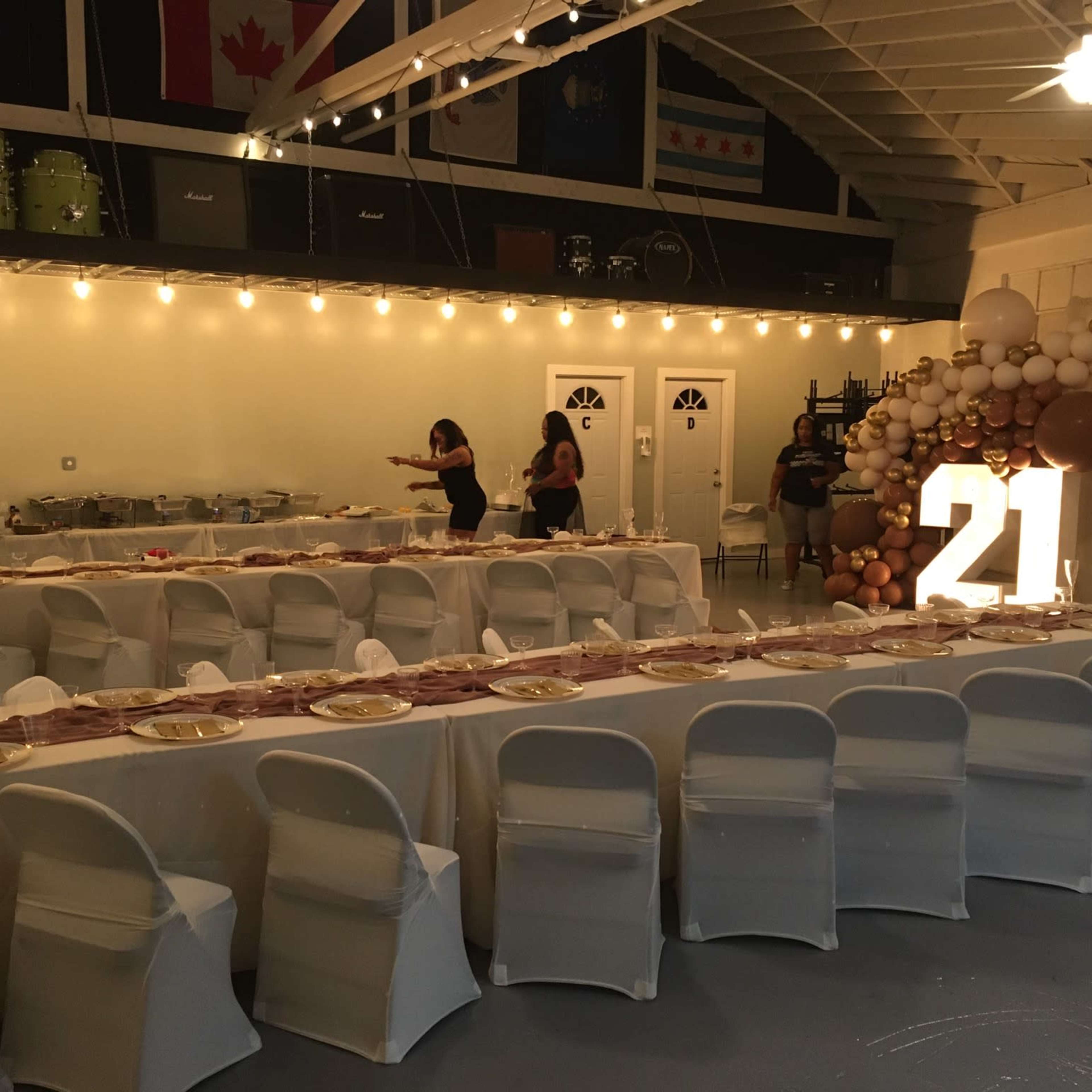 A decorated venue set up for a 21st birthday celebration, featuring a long dining table with plates and napkins, illuminated "21" numbers, and a backdrop of balloons and streamers.