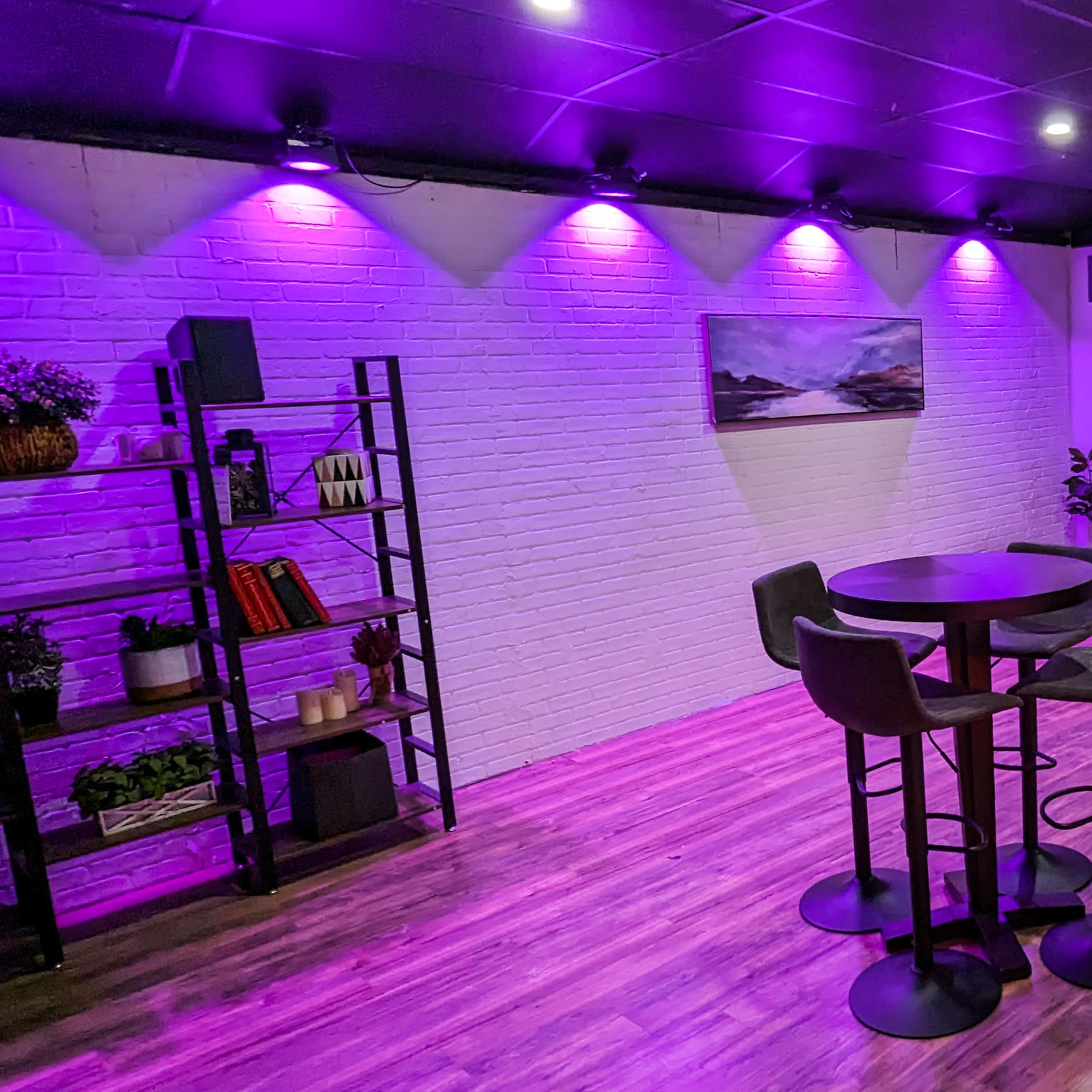 The room features a seating area with a gray couch, a round table surrounded by bar stools, and purple accent lighting illuminating the walls.