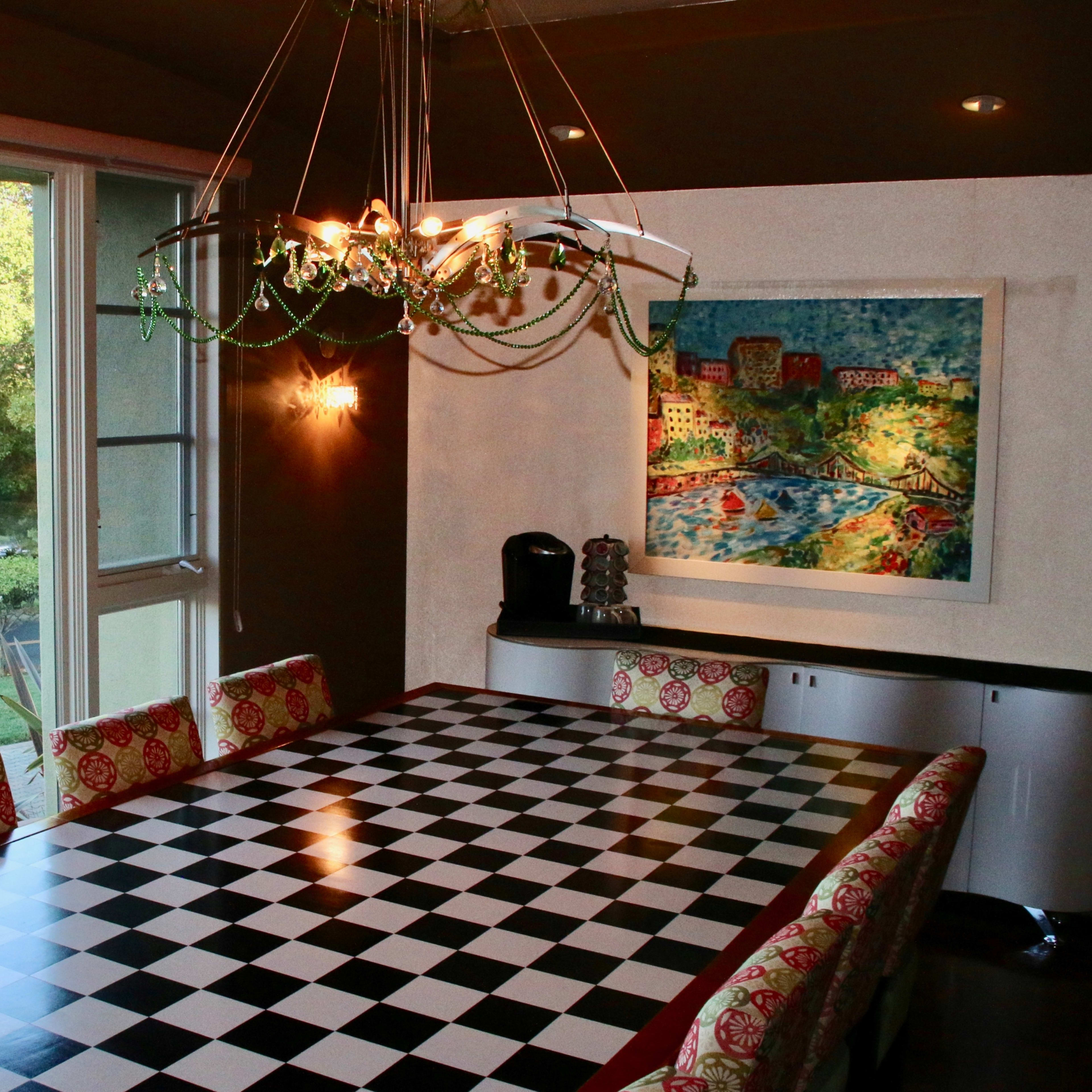 A large dining table with a checkered pattern is set in a well-lit room featuring a window that offers a view of greenery outside.