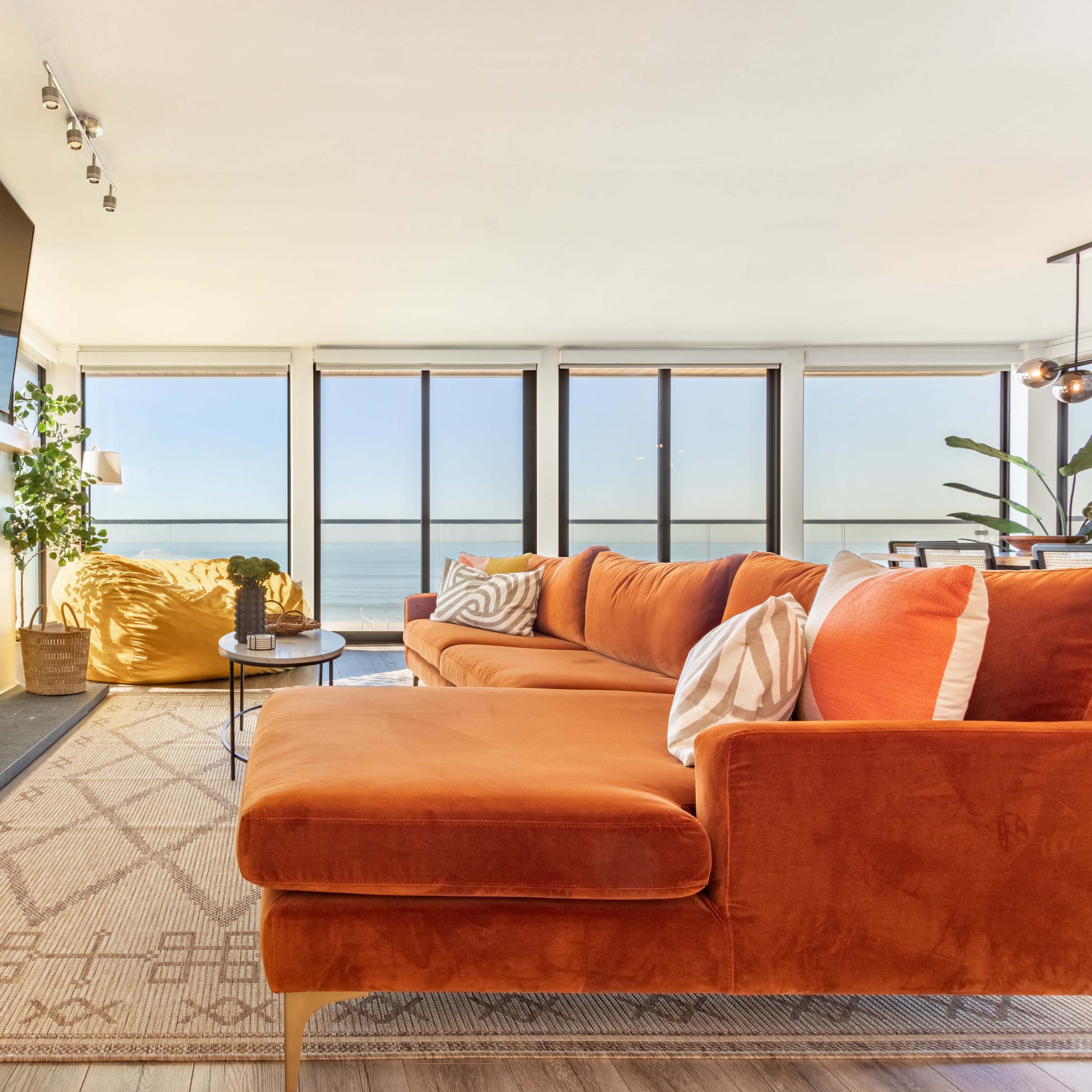 A modern living room features an orange sectional couch, a stone fireplace with a mounted television above it, and large windows overlooking a body of water.