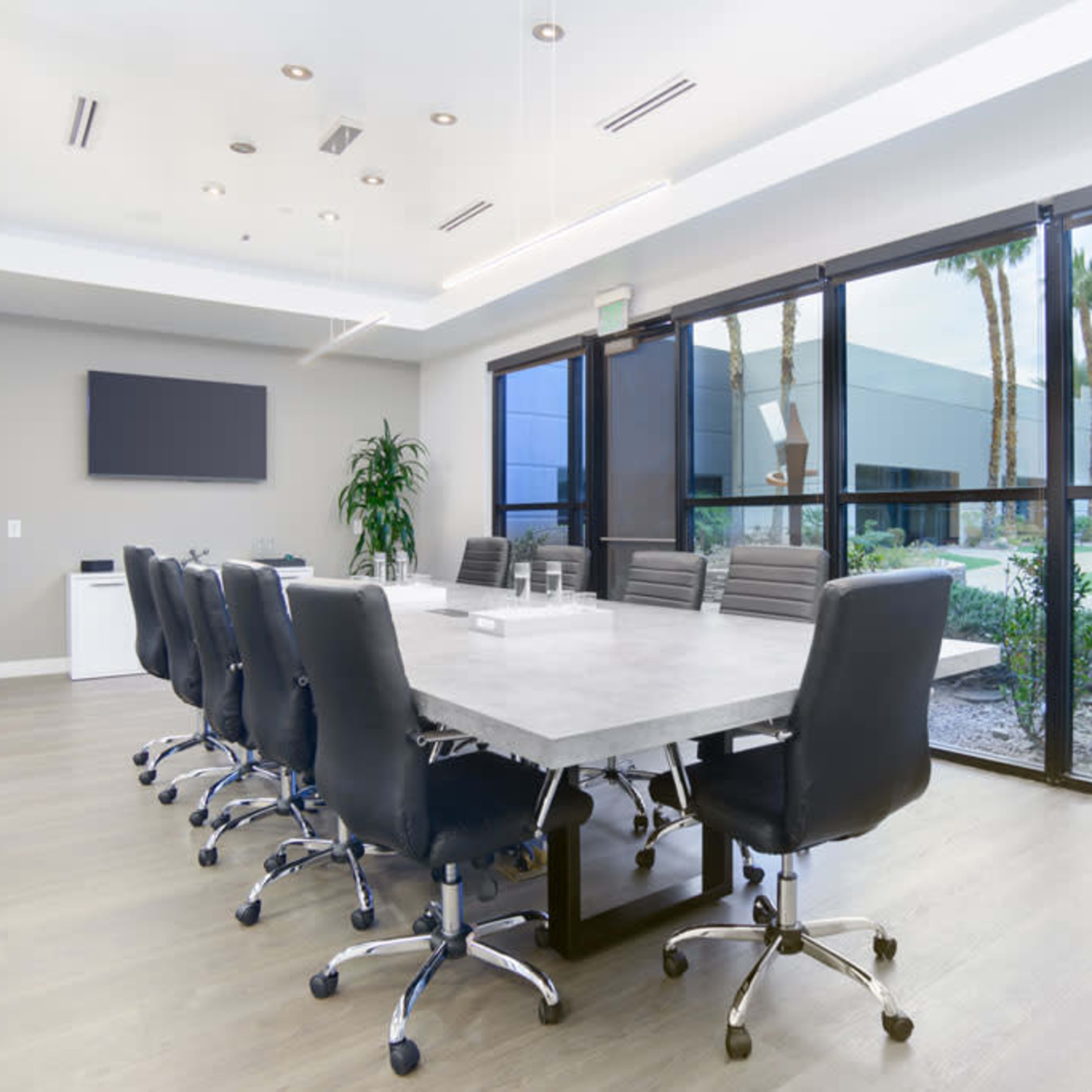 A modern conference room features a long table surrounded by black office chairs, with large windows overlooking a landscaped area.