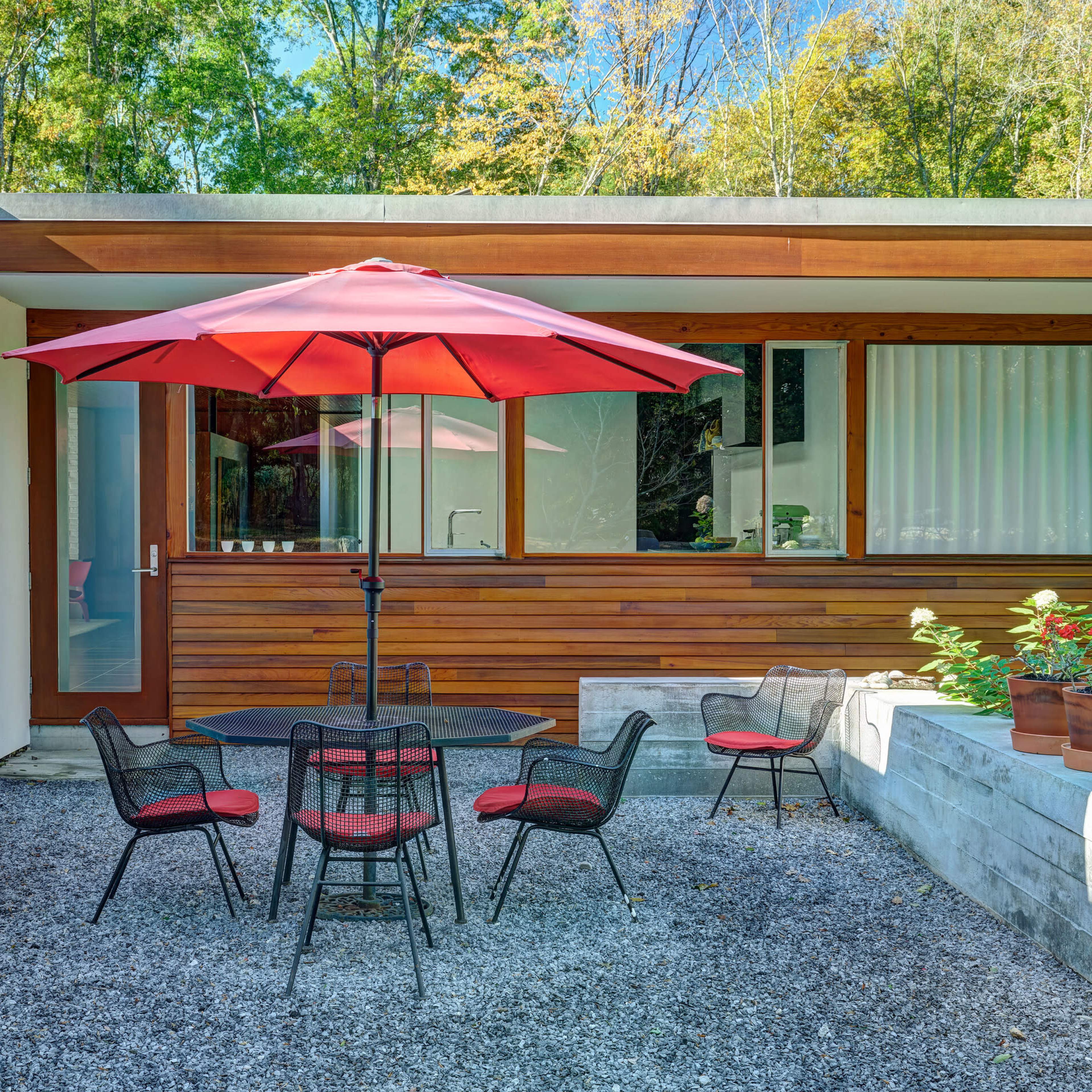 A patio area features a round table surrounded by black chairs with red cushions, topped by a large red umbrella, alongside a modern wooden and glass home.