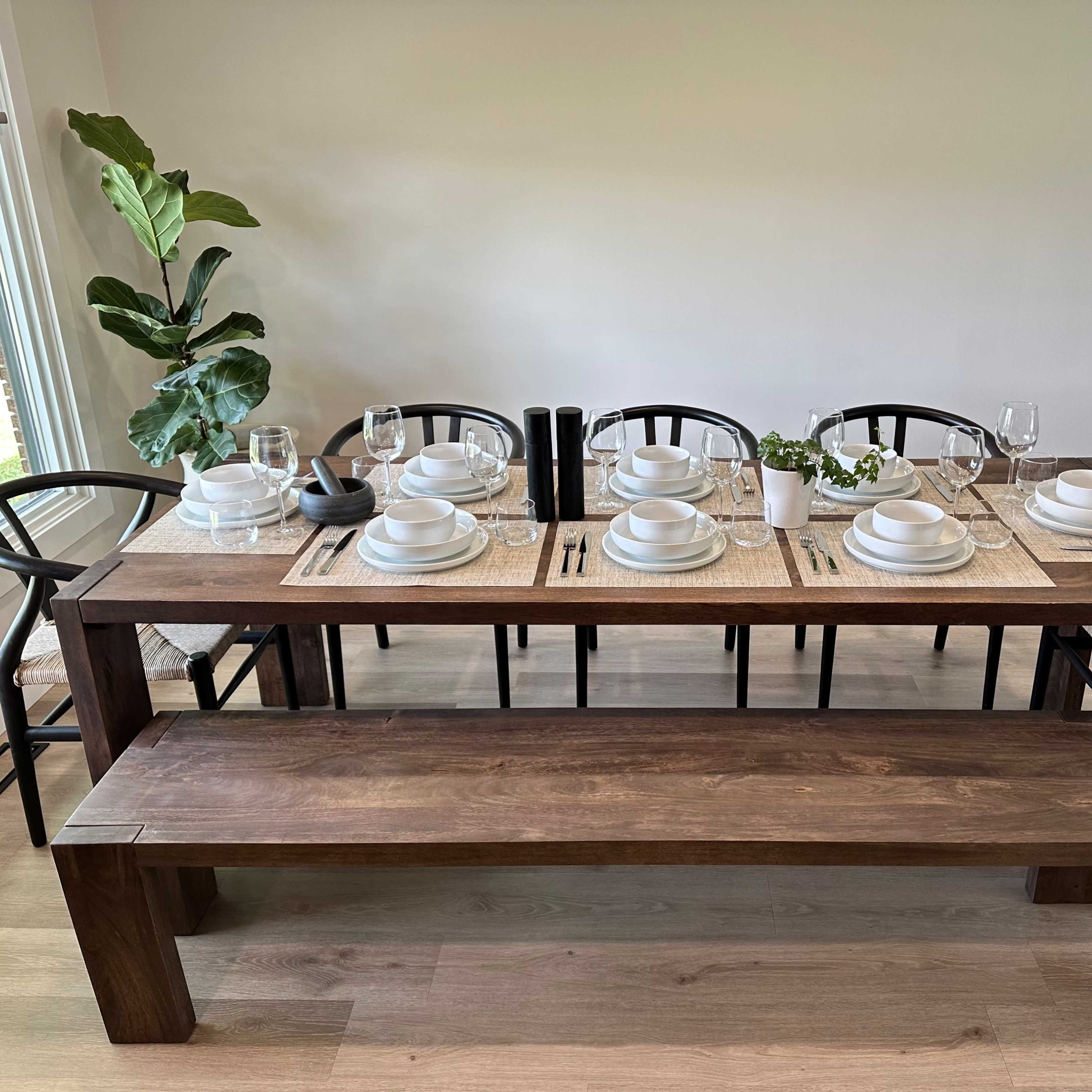 A wooden dining table is set with plates, glasses, and utensils, accompanied by a potted plant and a bench, in a well-lit room.