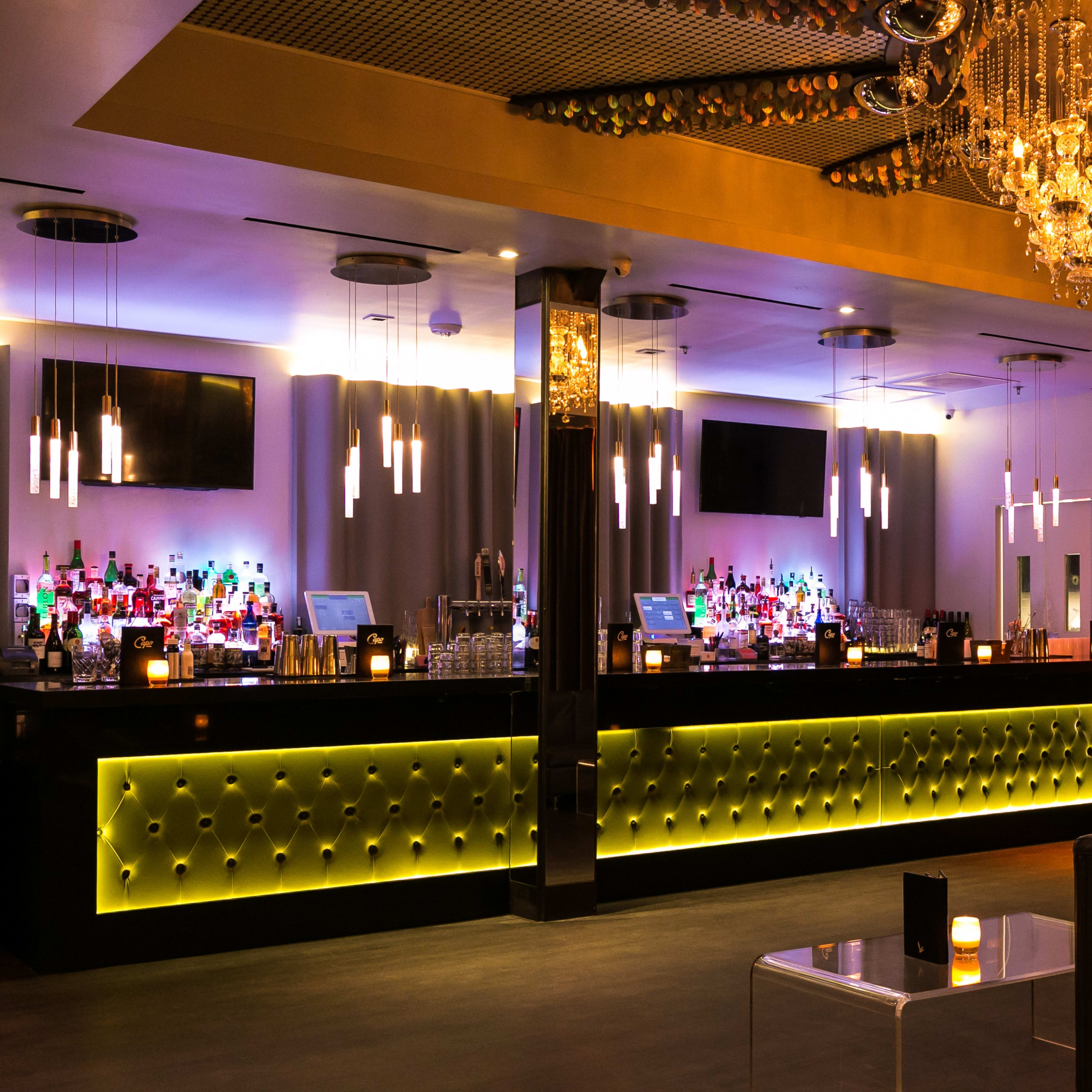 A stylish bar interior with a brightly lit counter, shelves of colorful bottles, and a sparkling chandelier overhead.