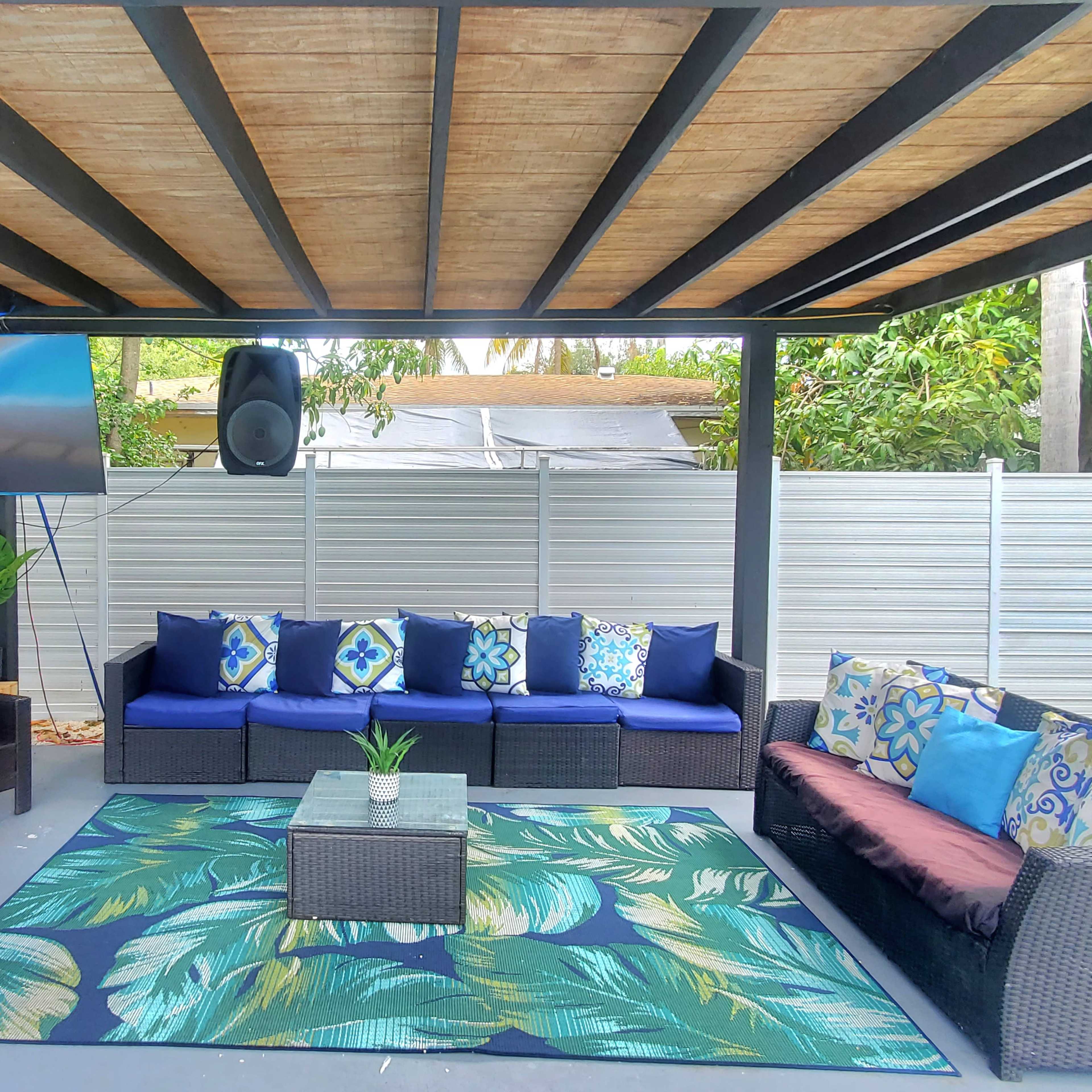 A covered outdoor patio features dark wicker furniture with colorful cushions arranged around a large area rug, along with a television mounted on the wall.