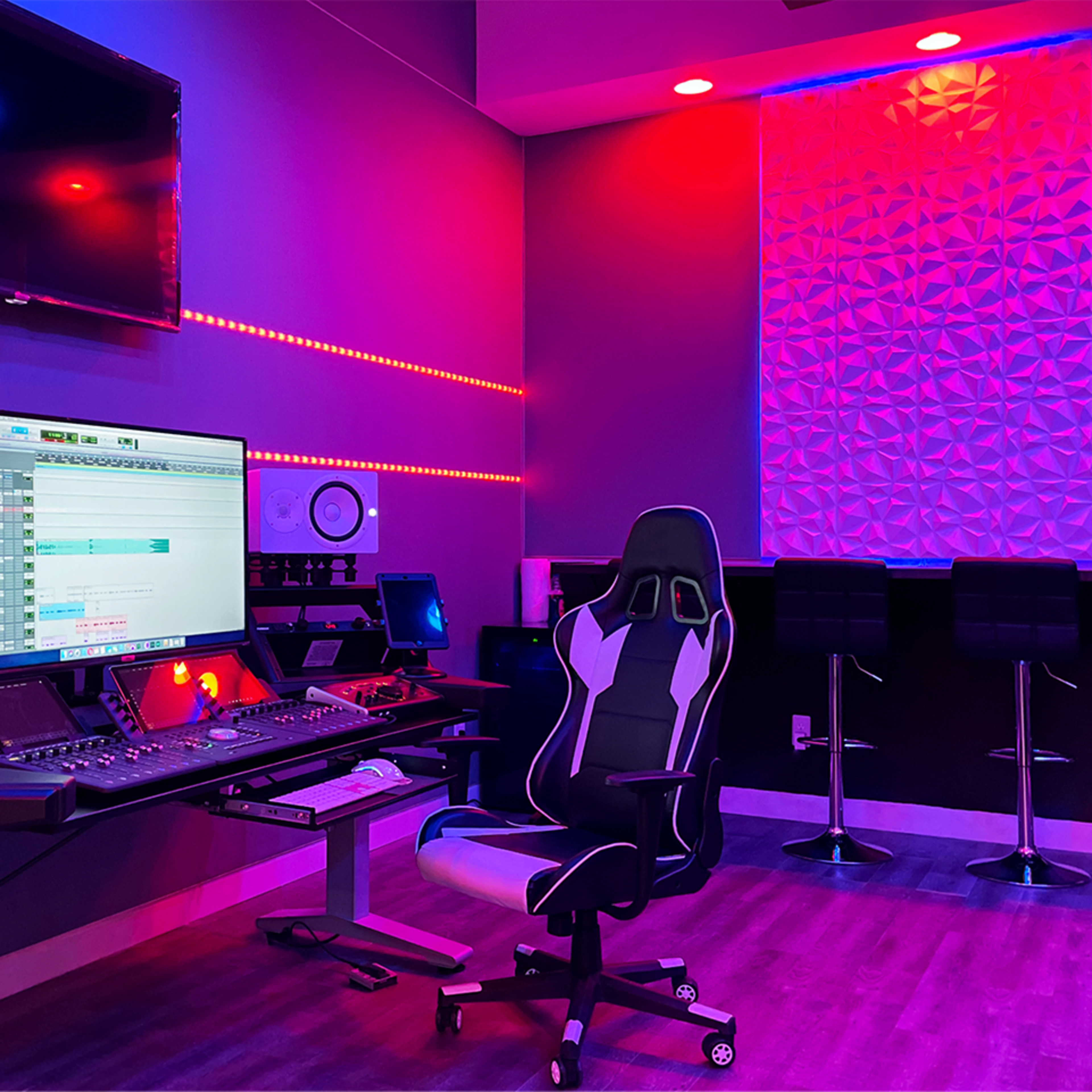 A modern recording studio features a large computer monitor, a sleek mixing desk with audio equipment, and colorful LED lighting.