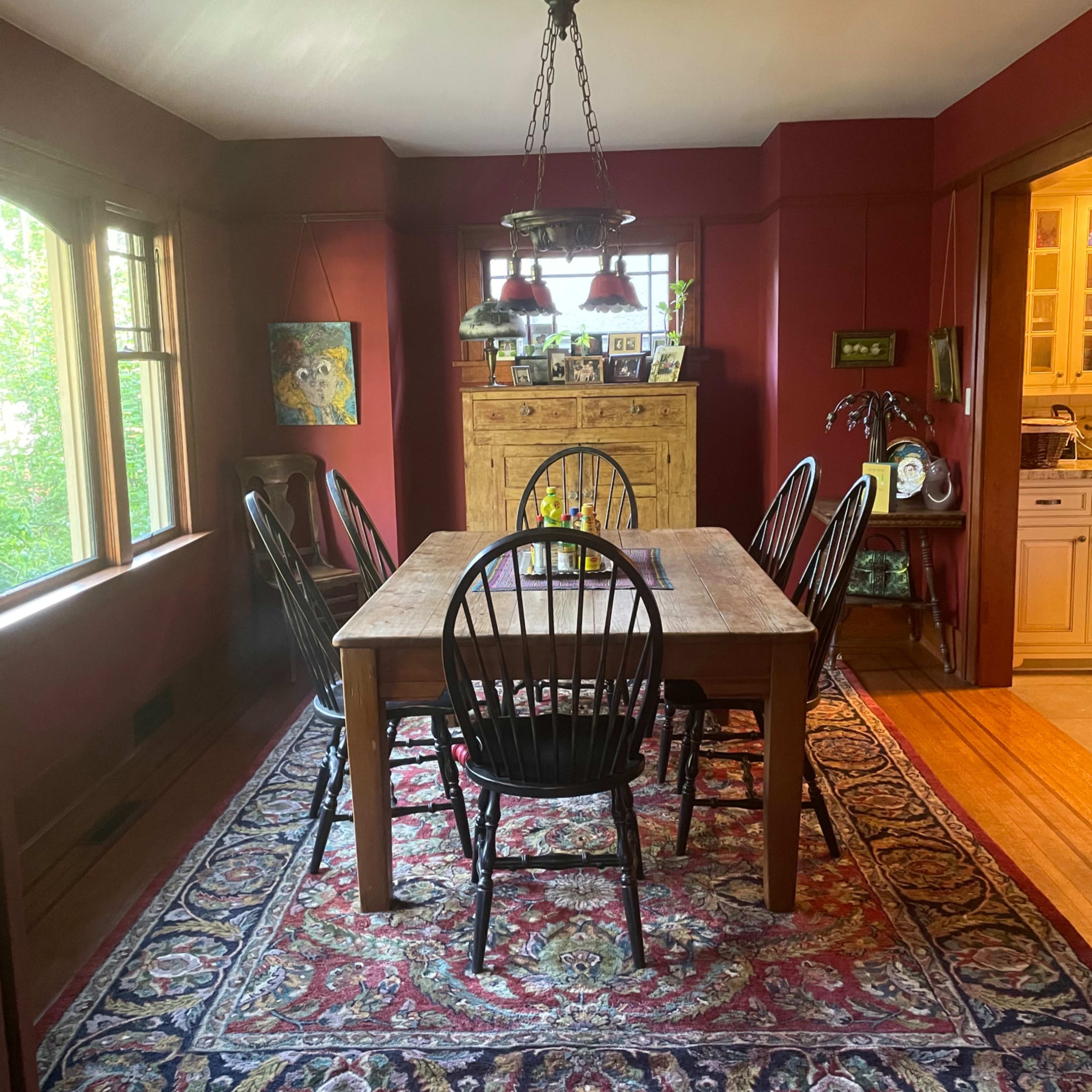 A dining room features a wooden table surrounded by black chairs, with large windows and a patterned rug on the floor.