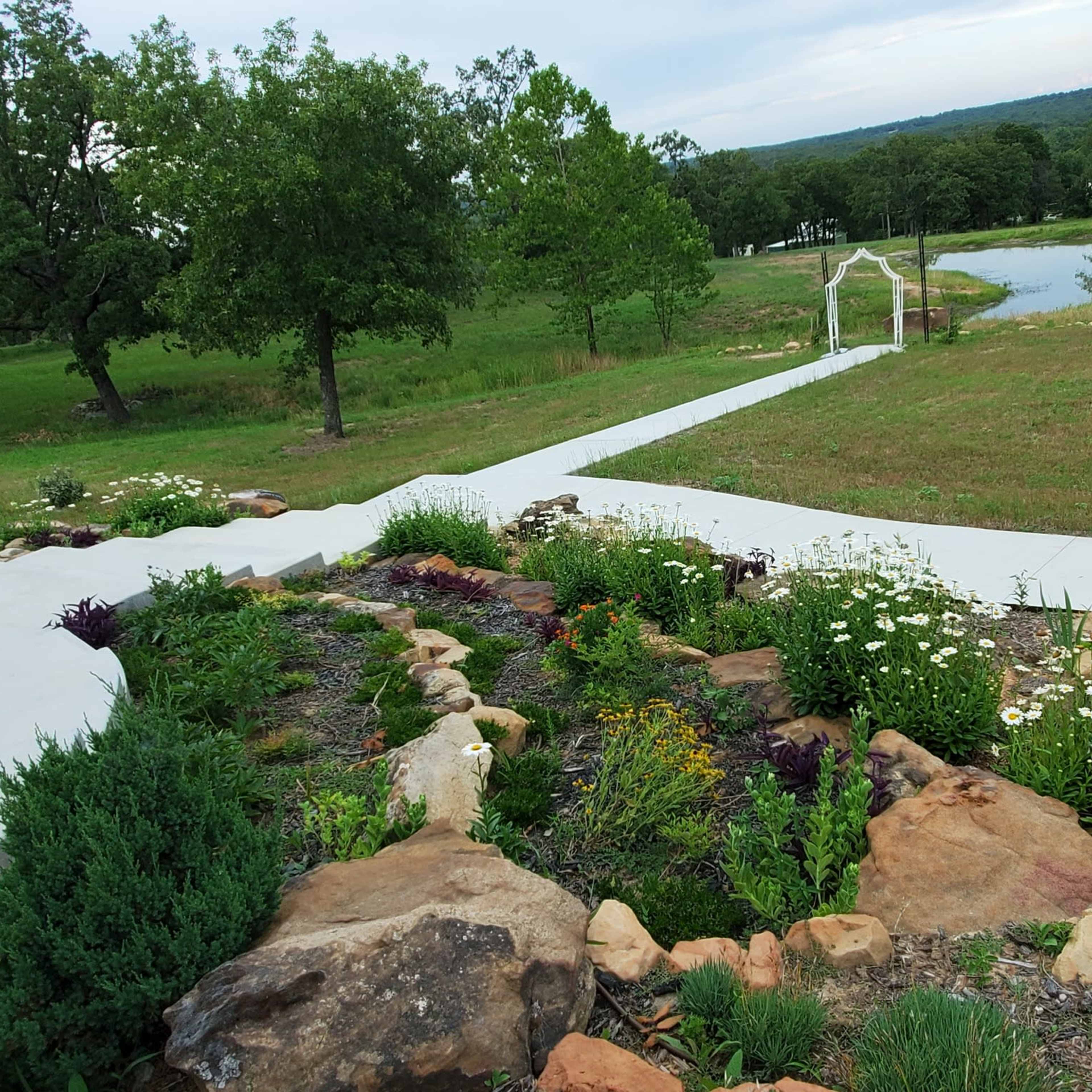 A pathway lined with colorful flowers leads down a slope toward a pond and a white archway under a blue sky.