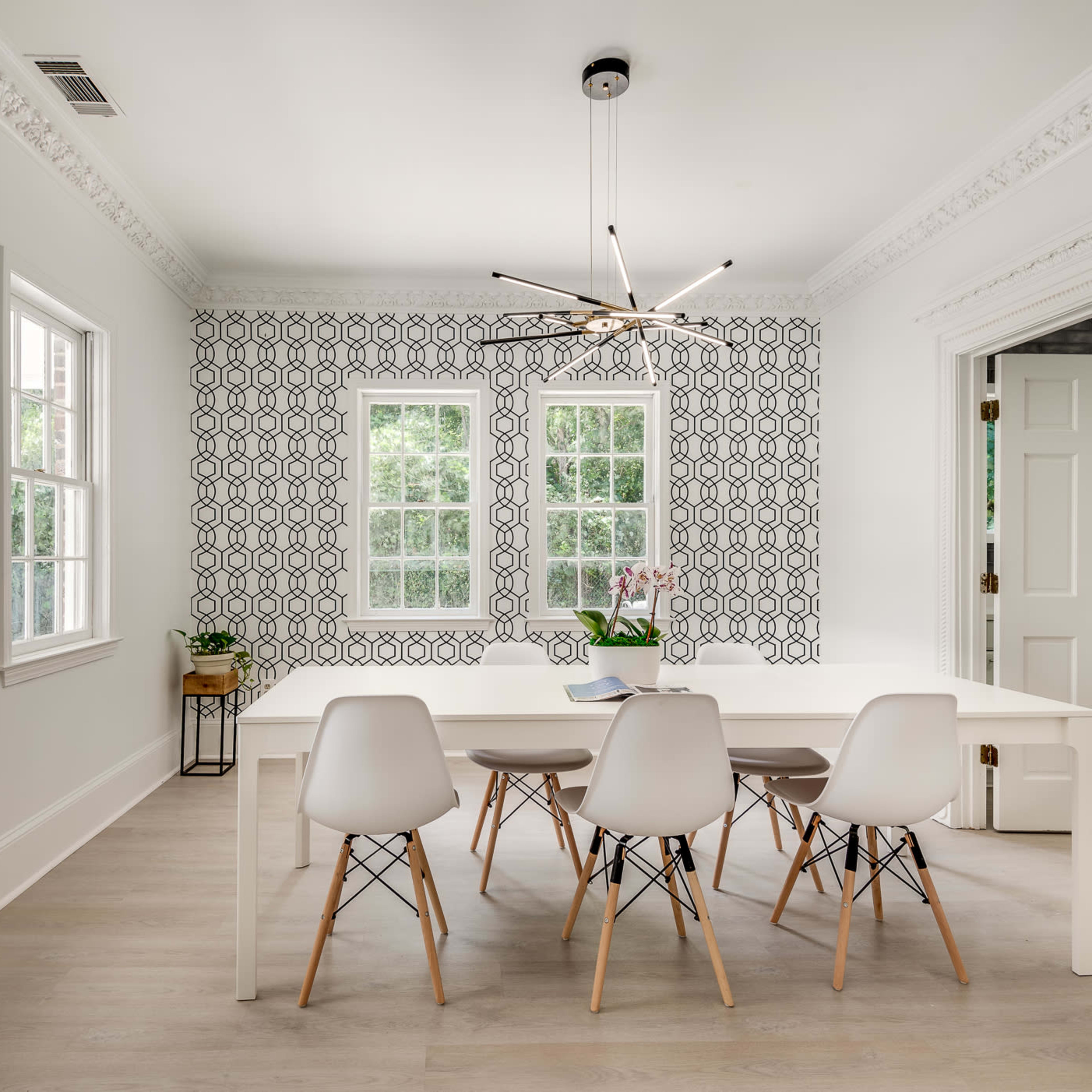 A bright dining room features a white table with six chairs, surrounded by patterned wallpaper and large windows.