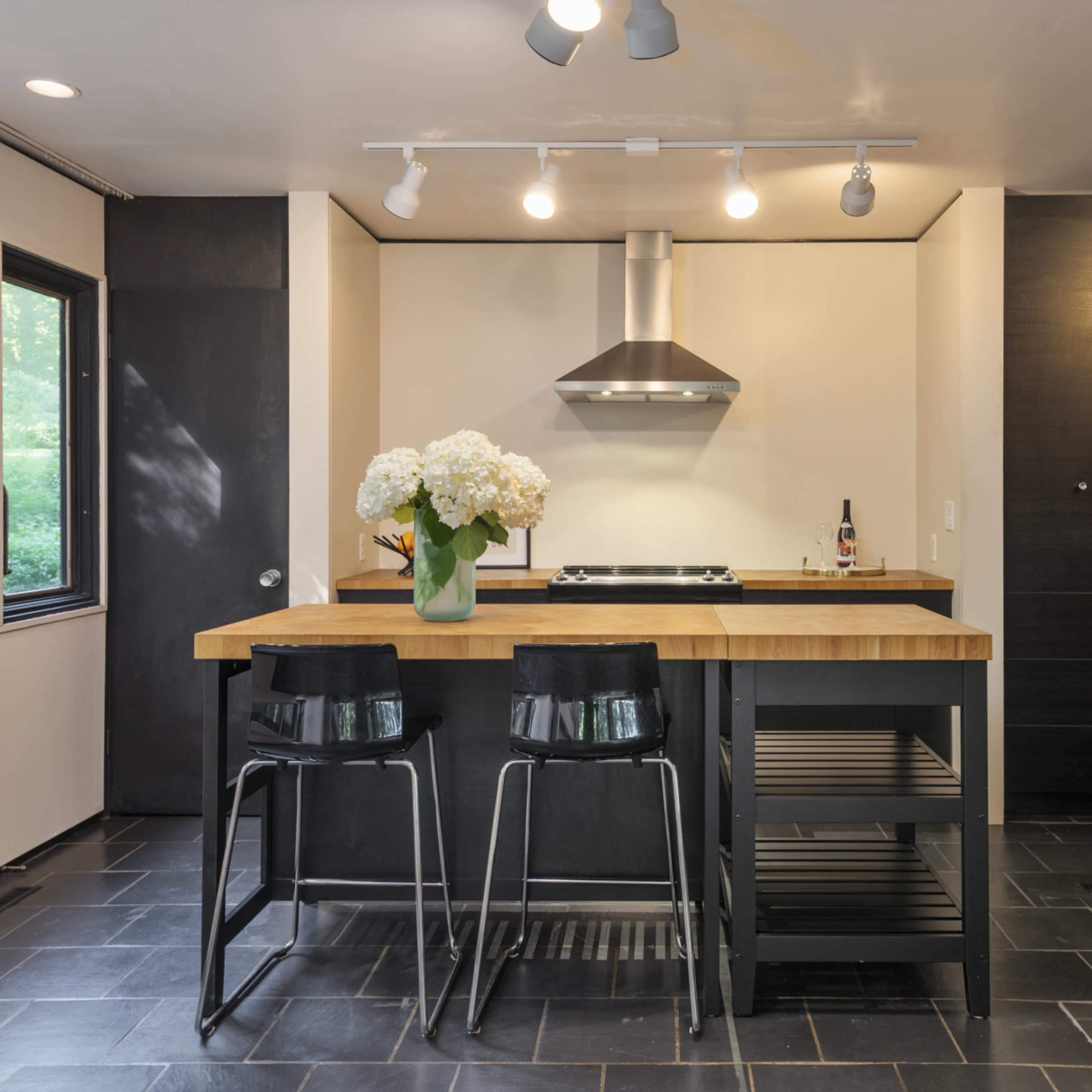 A modern kitchen featuring a central island with two black stools, stainless steel appliances, and large windows displaying greenery outside.