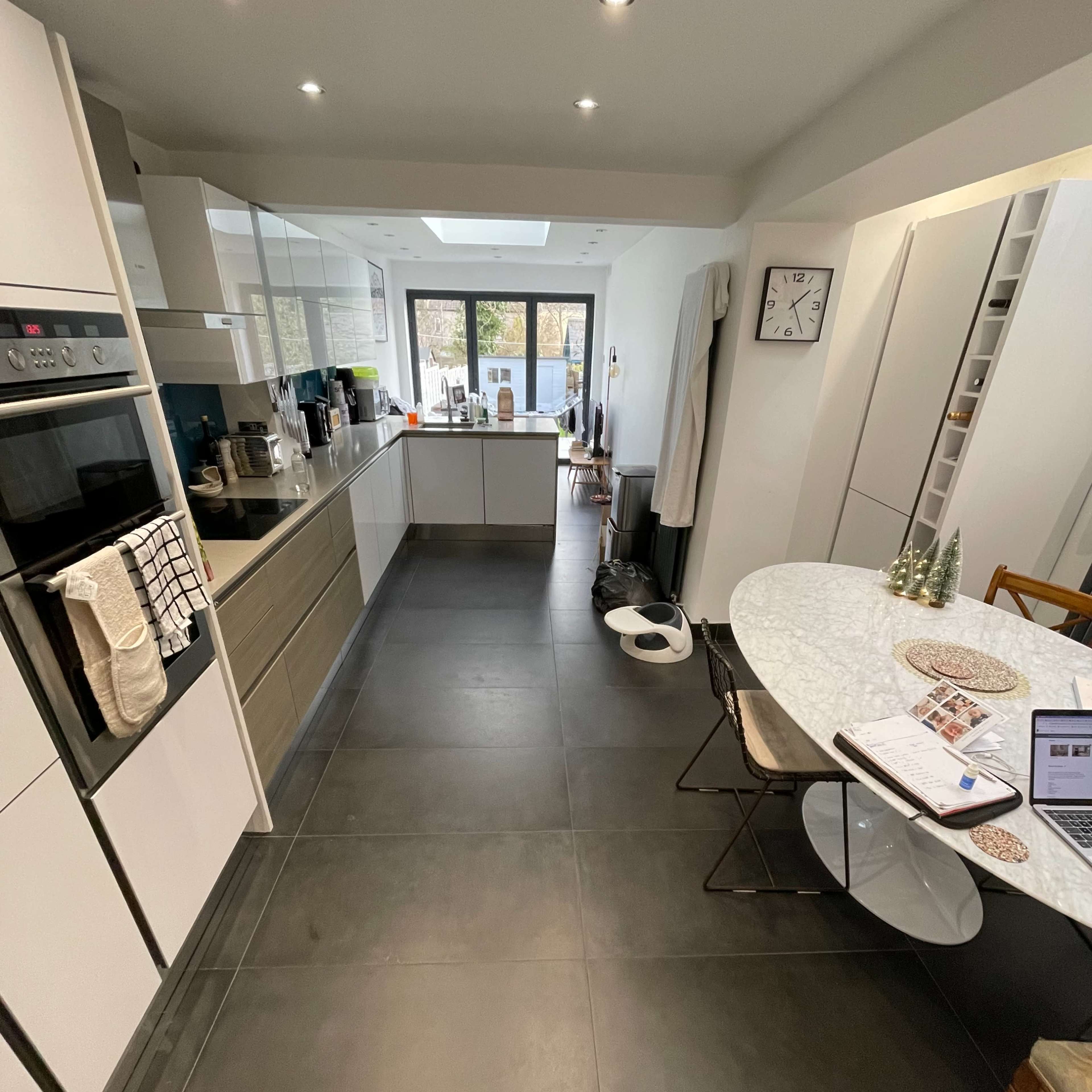 A modern kitchen featuring a long, narrow layout with white cabinets, a circular dining table, and large windows allowing natural light.