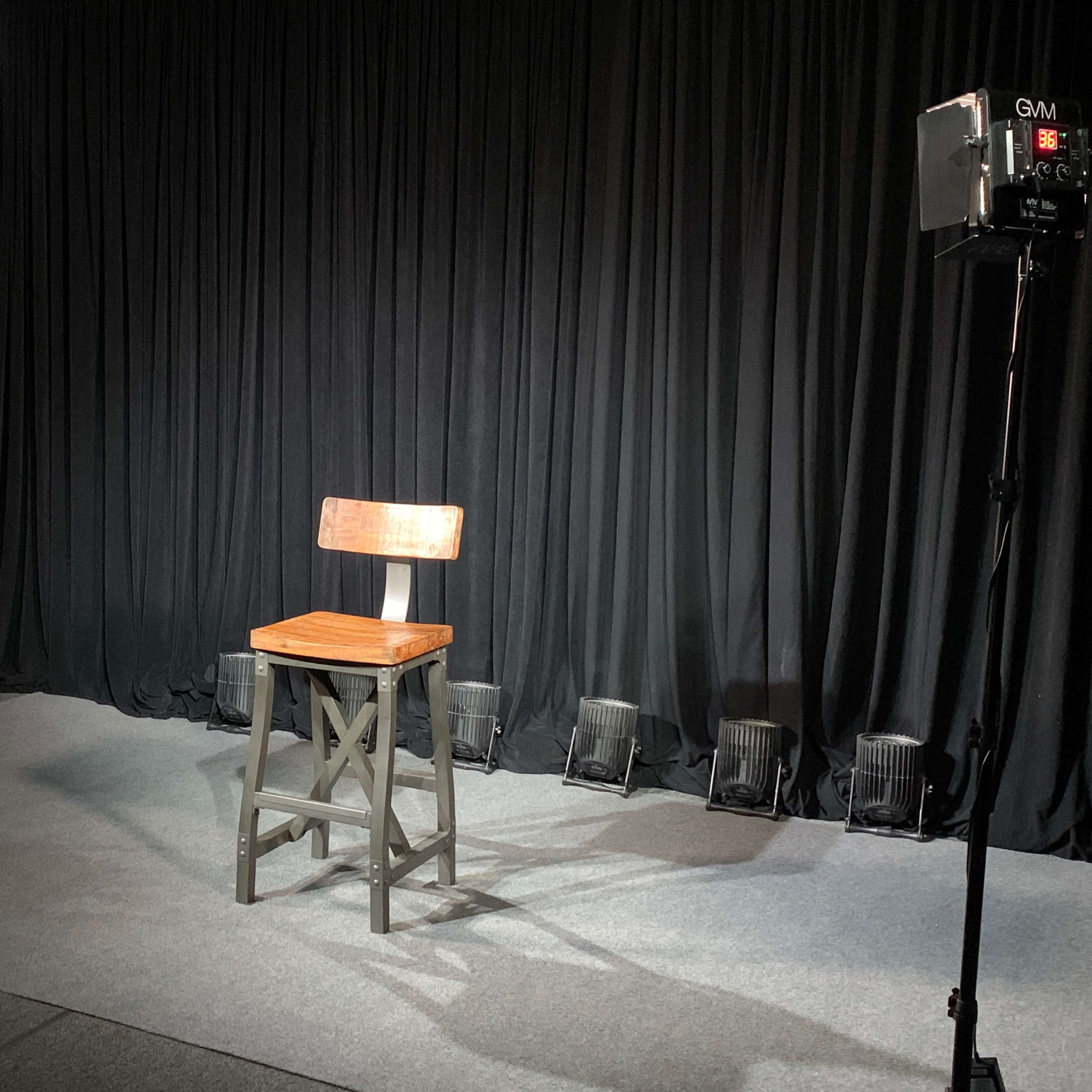 A wooden stool is positioned on a gray carpeted area, surrounded by two lights and a backdrop of black curtains.